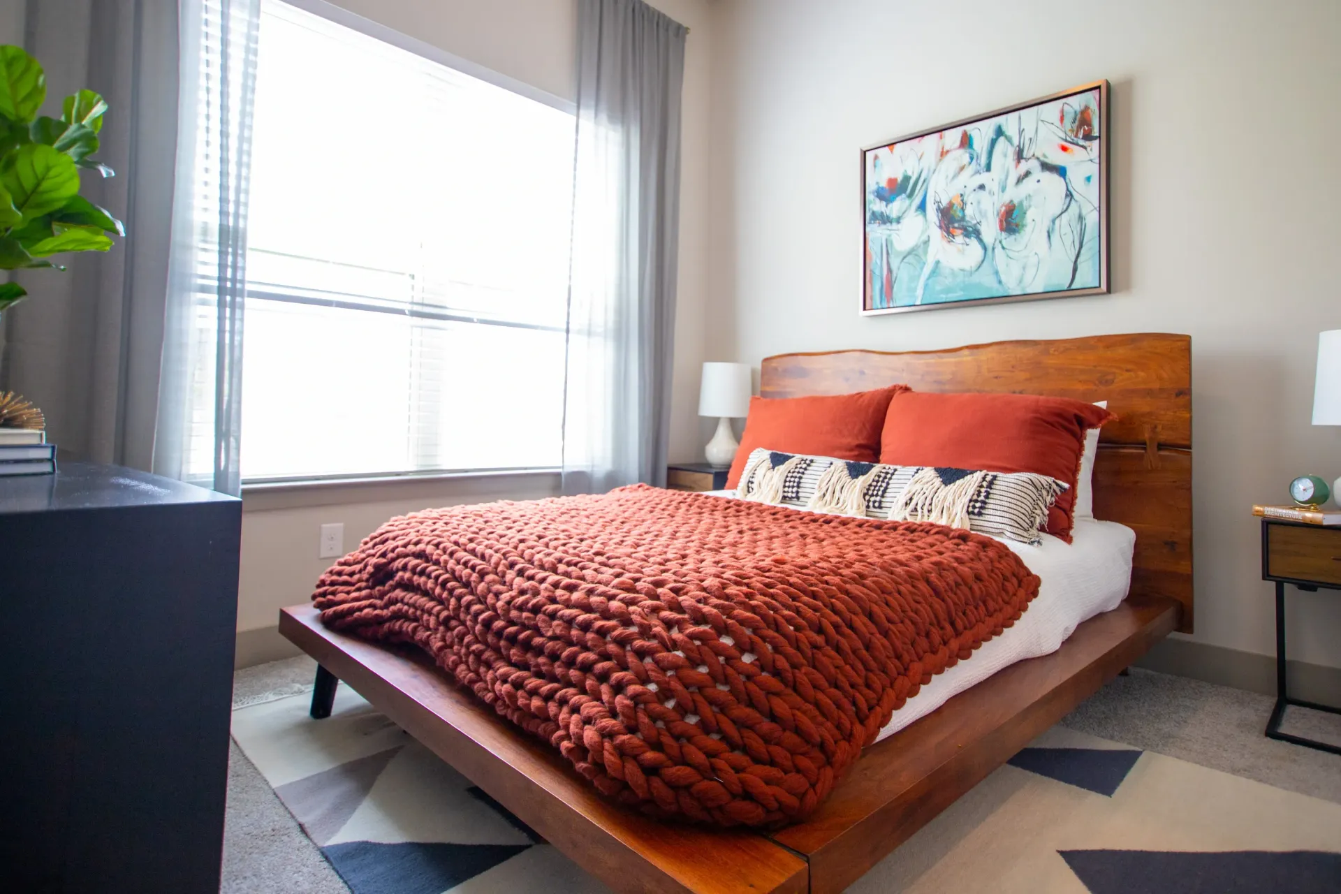 Bedroom with wooden platform bed, rust knit blanket, pillows, plant, and a large window with sheer curtains.