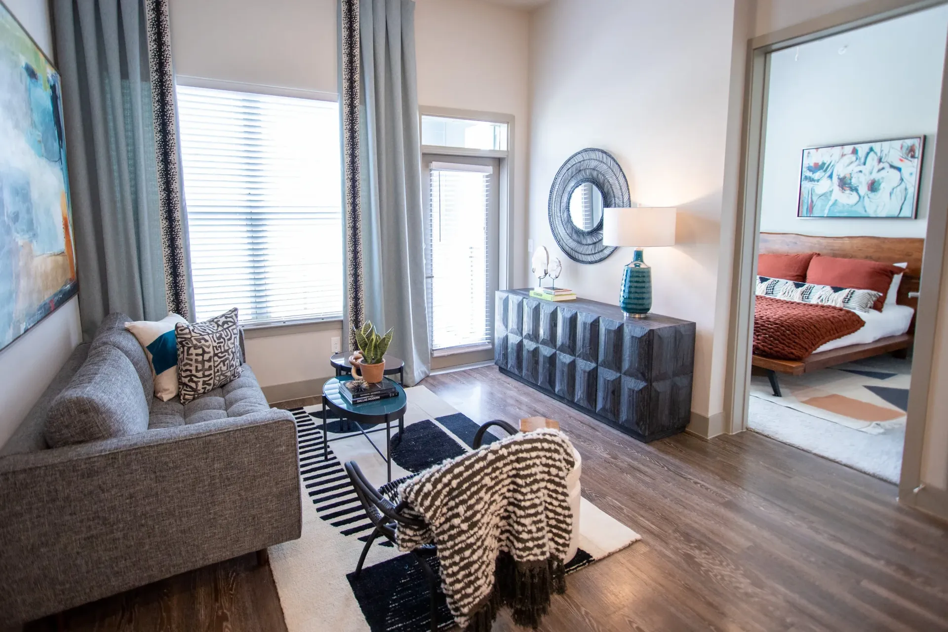 Living room in an apartment: gray sofa, coffee table, rug, and a textured dresser by large windows.