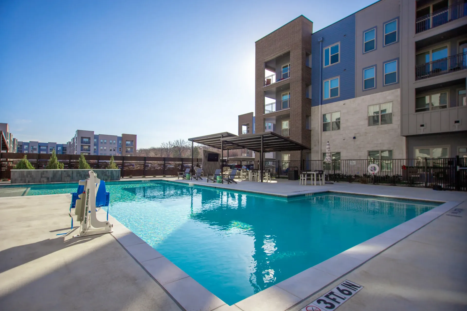 Outdoor apartment community pool with lounge chairs and shaded seating area beside modern buildings.