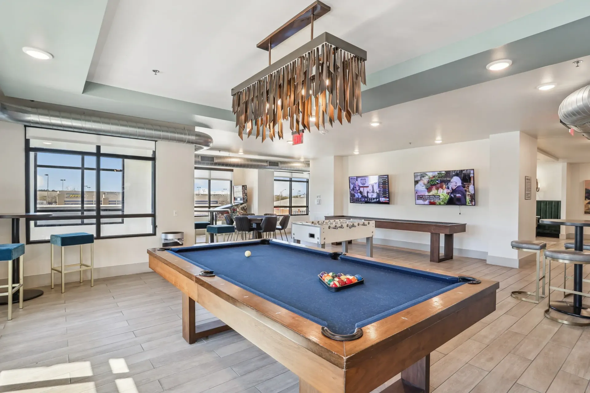 Indoor community recreation room with a pool table, foosball, TVs, and seating by large windows.
