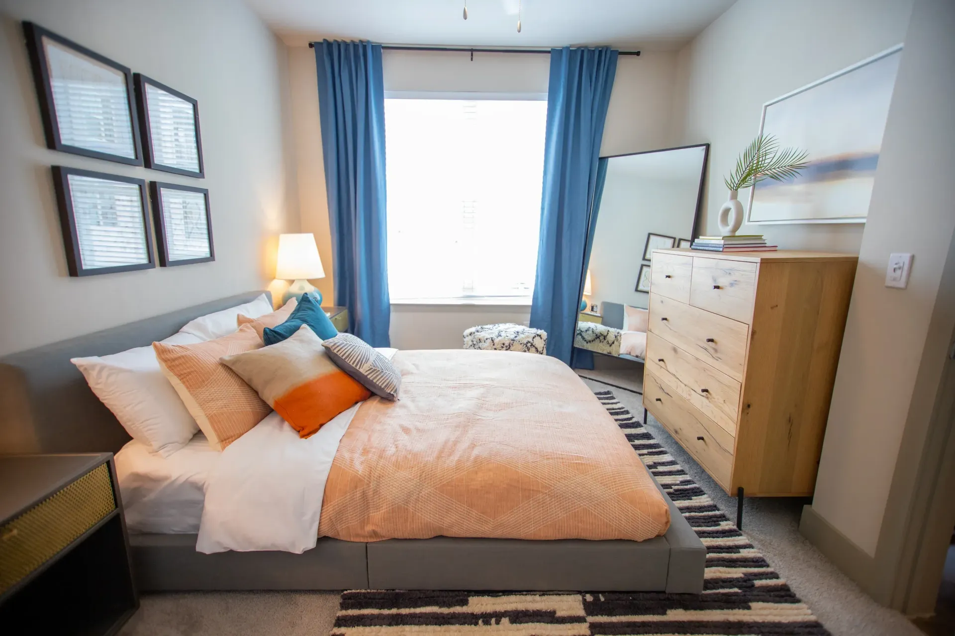 Bedroom in a modern apartment with a low-profile bed, peach bedding, blue curtains, and a wooden dresser.