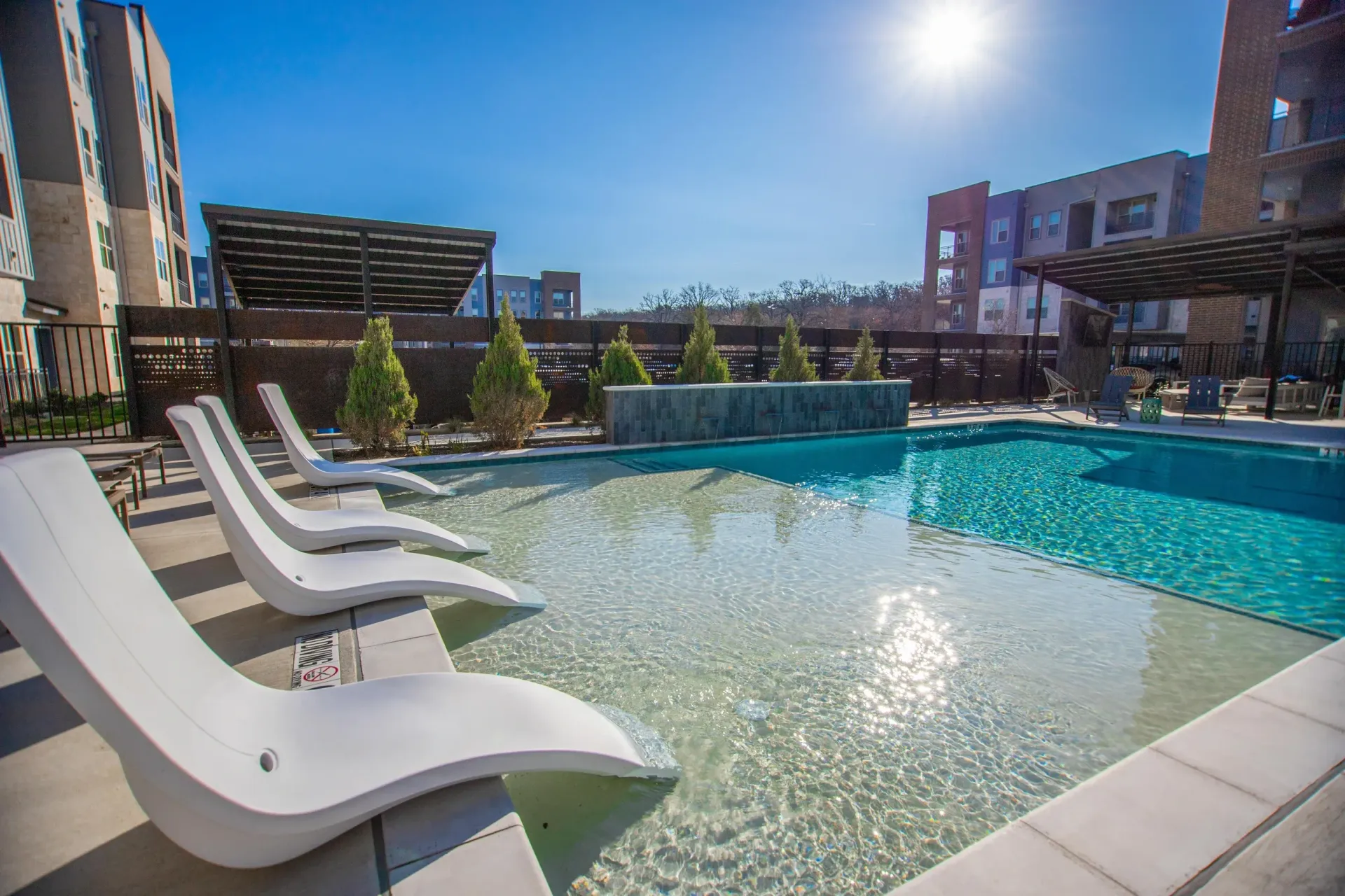 Outdoor pool area with white lounge chairs and clear water among modern apartment buildings.