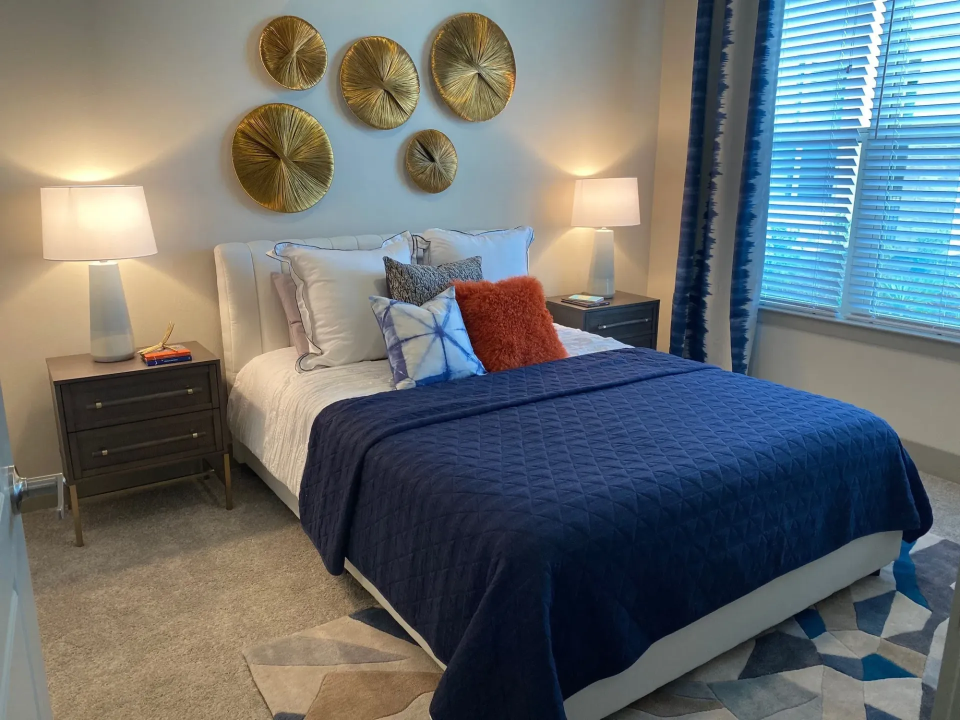 Bedroom in apartment with white bed, blue quilt, pillows, nightstands, lamps, and wall decor.
