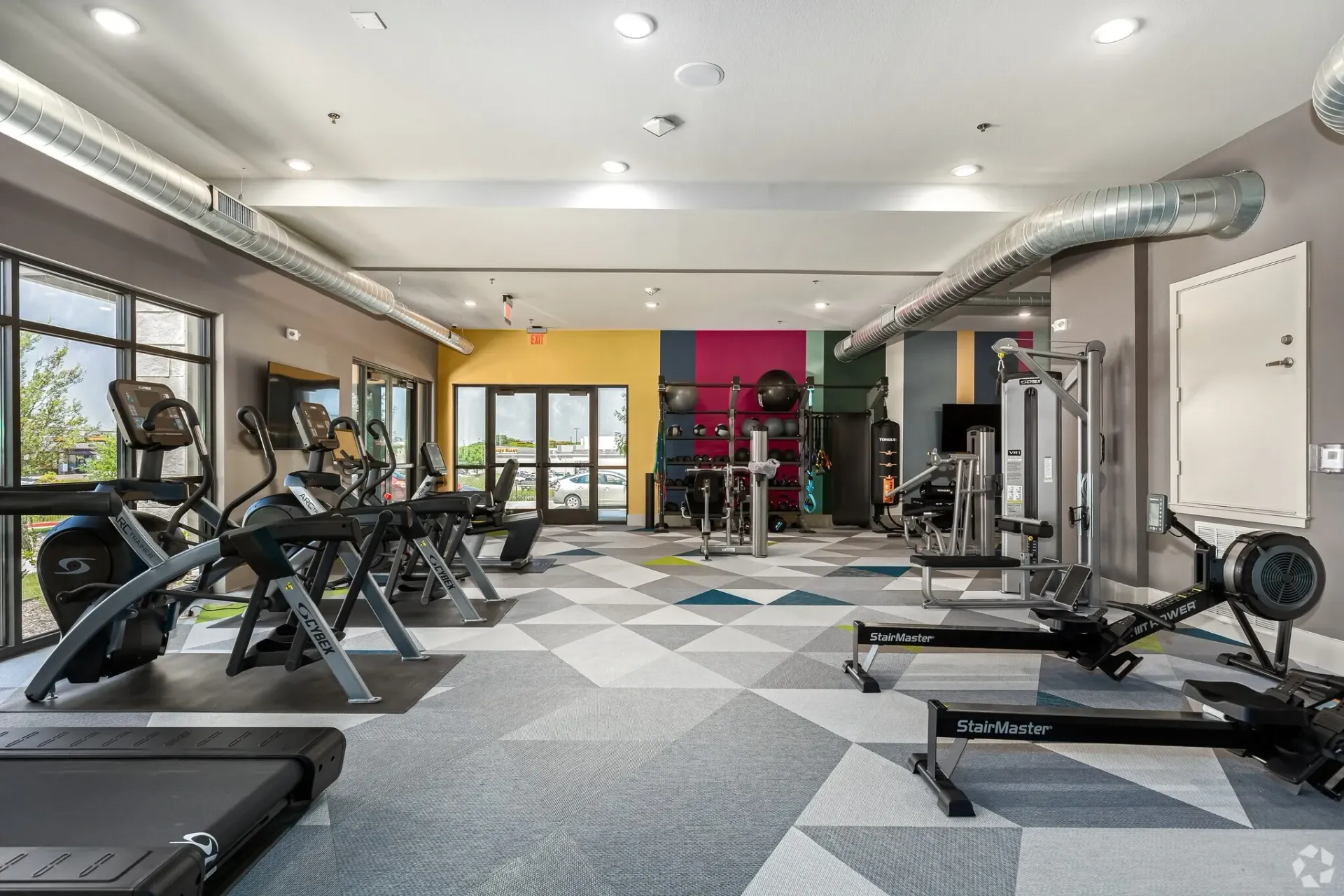 Interior community gym with cardio machines, weight rack, and colorful wall accents.