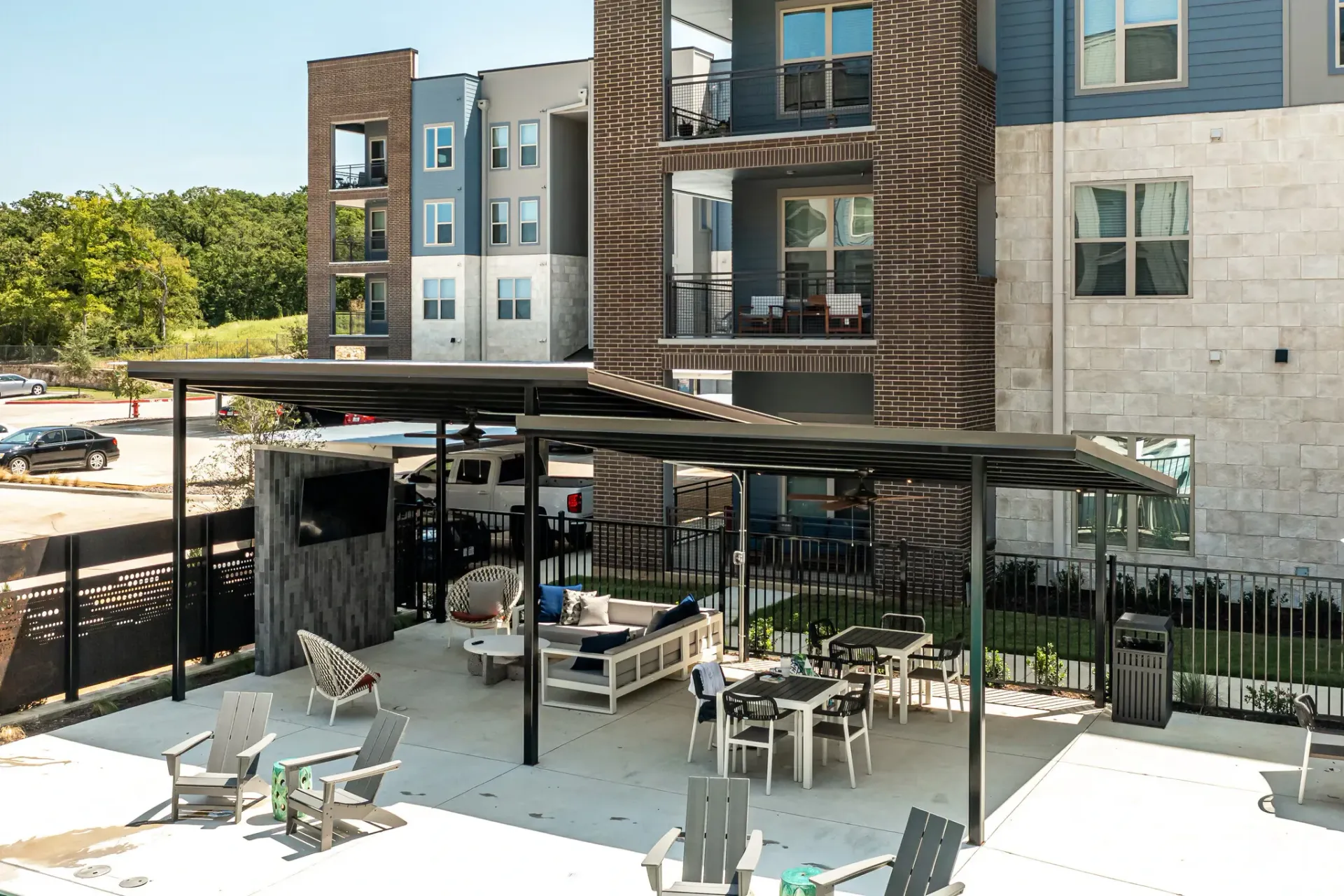 Outdoor communal lounge with couches, chairs, and tables under a covered pavilion beside apartment buildings.