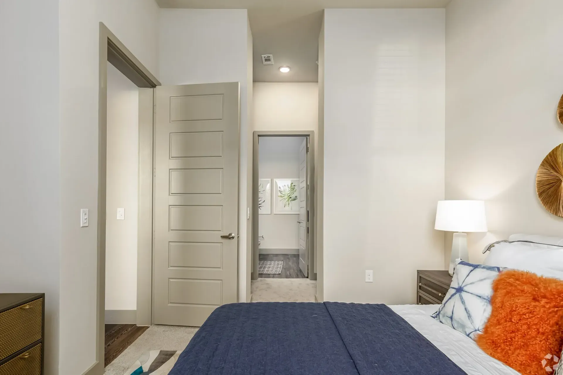 Bedroom in a modern apartment with a bed, nightstand, lamp, and doorway to a hallway.