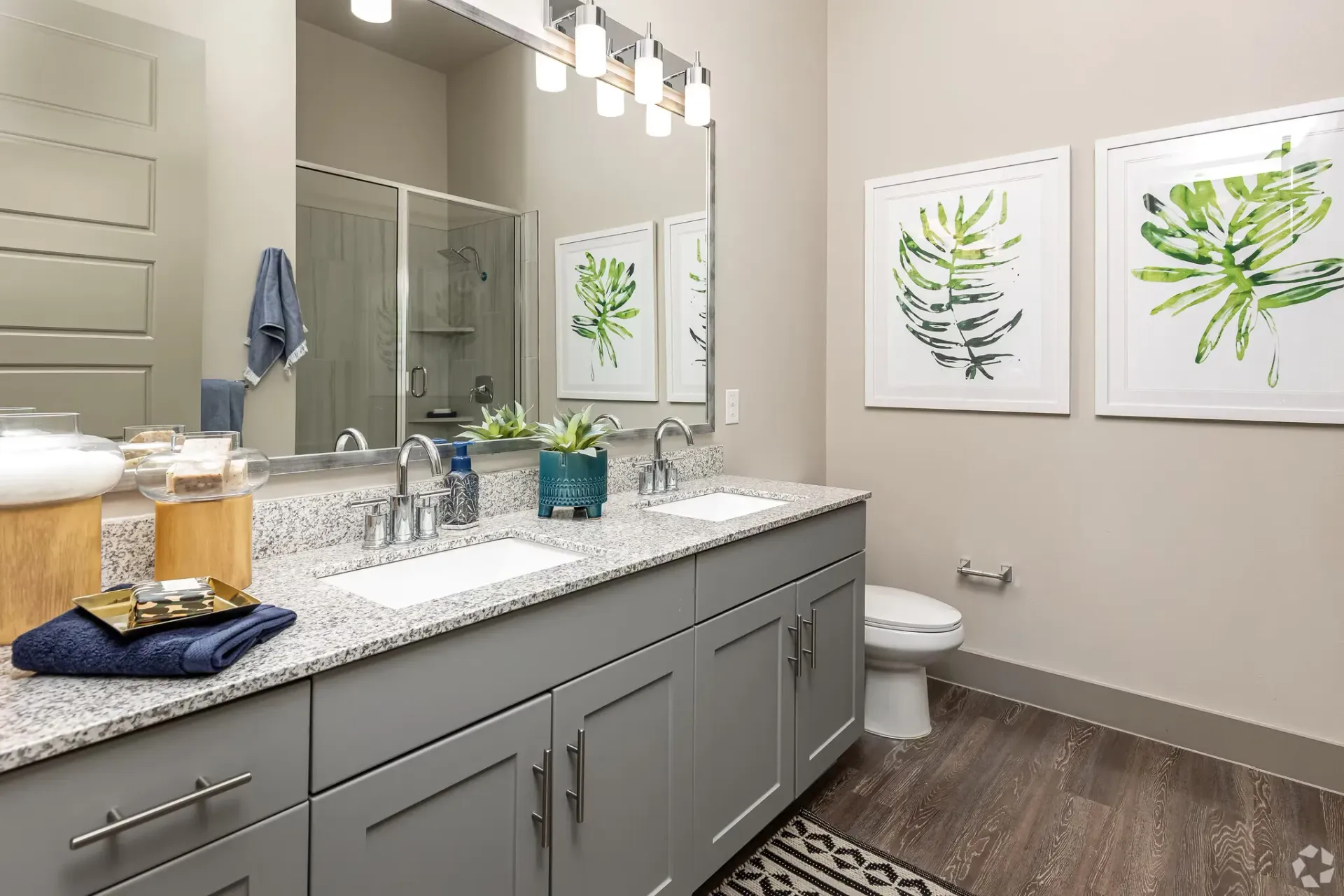 Bathroom with a double-sink vanity, granite countertop, gray cabinets, large mirror, and a glass-enclosed shower.