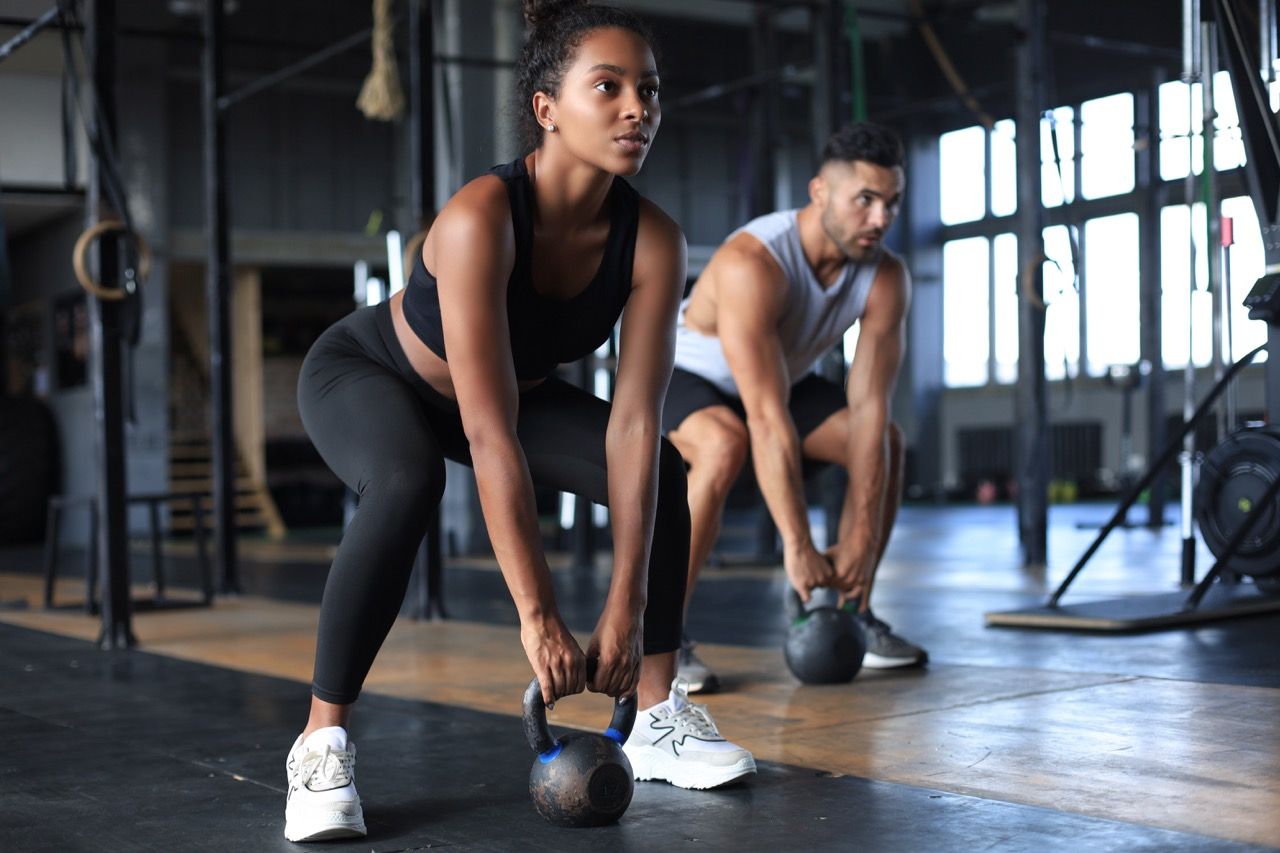 Two people doing kettlebell deadlifts in a gym, focused on training on a wooden floor.