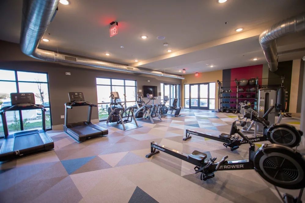 An indoor gym featuring treadmills, rowers, and exercise bikes on a patterned floor with exposed ceiling ducts.