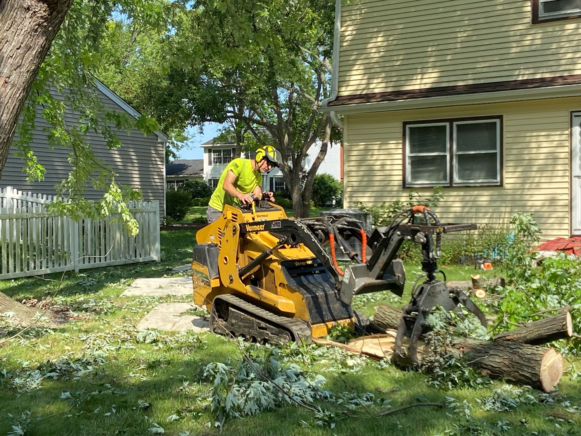 A worker uses a yellow mini-skid steer to grind a tree stump in a yard next to a house.