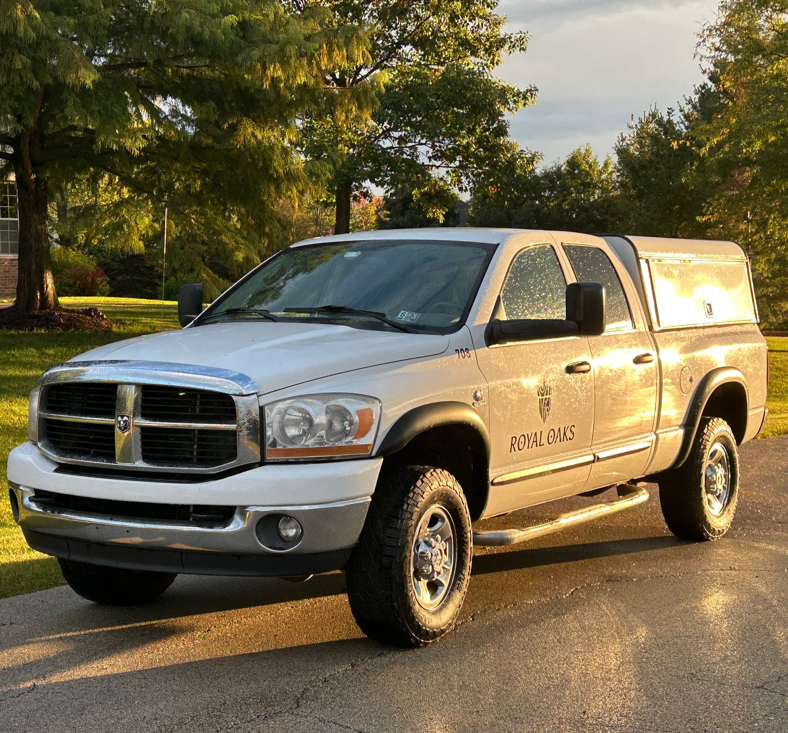A white Dodge Ram pickup truck with a camper shell parked on a paved driveway in front of trees.