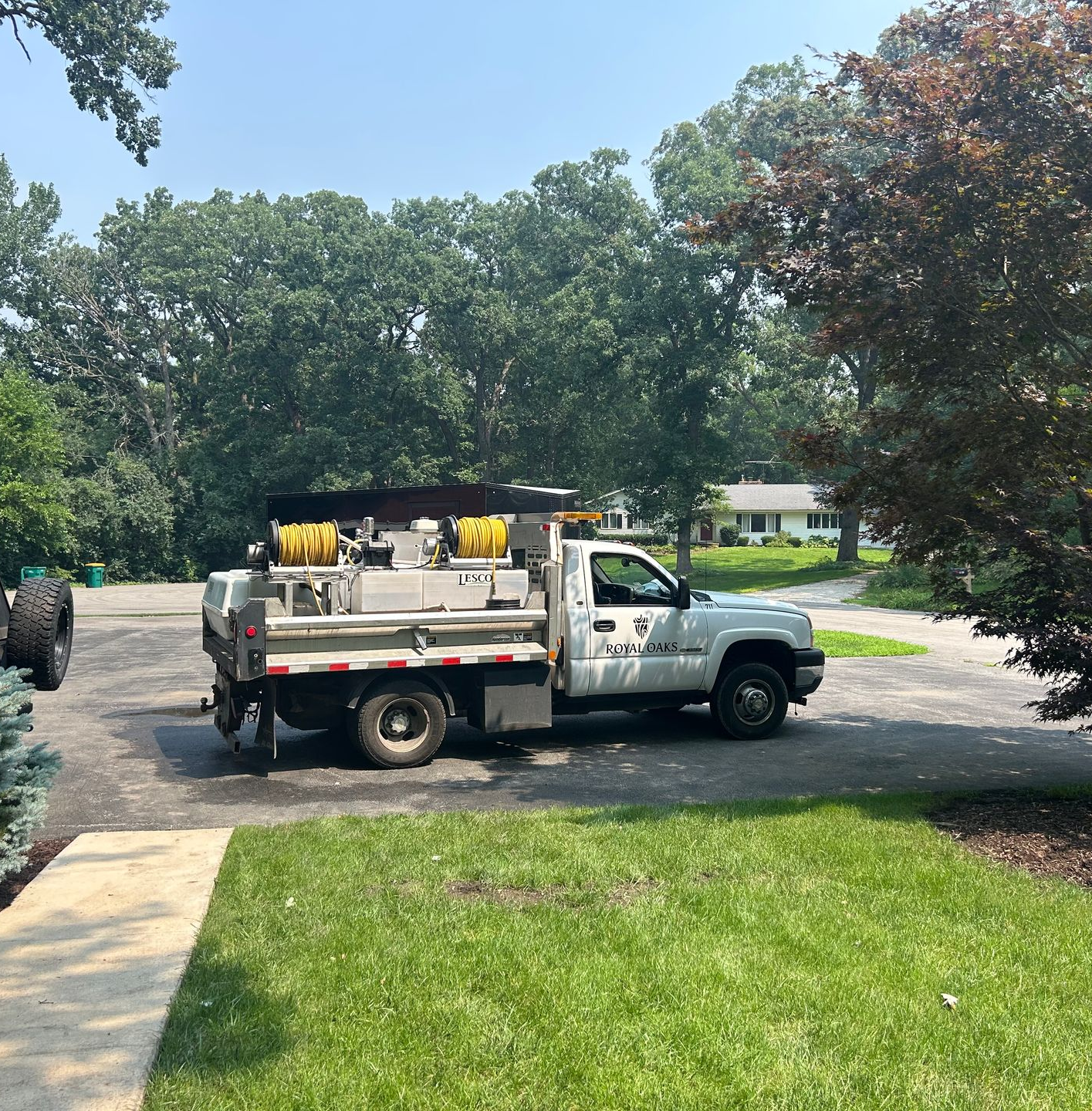 A white utility truck with two yellow hose reels parked on a paved driveway in front of a house and trees.