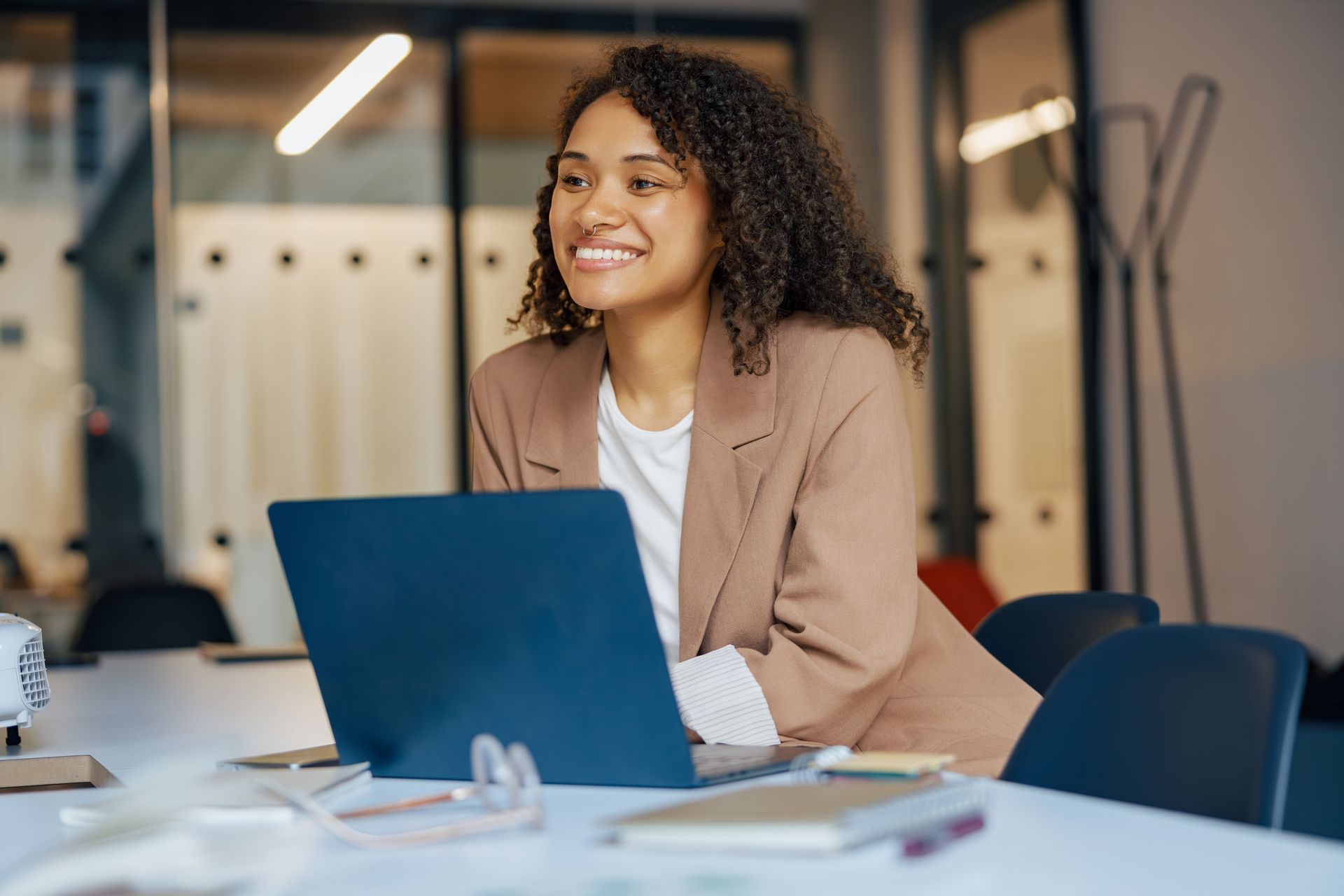 Smiling person in a tan blazer using a blue laptop at a desk in a modern office