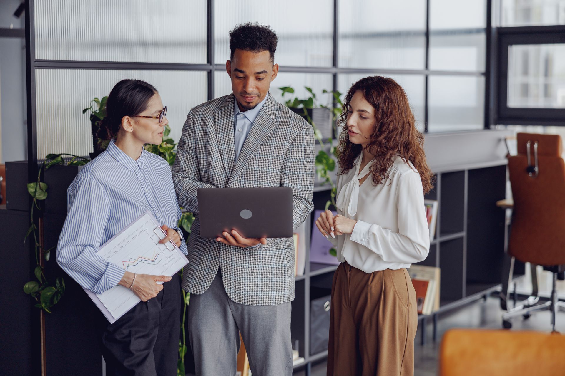 Three coworkers discussing a laptop in a modern office, with one holding papers and another holding a mug.