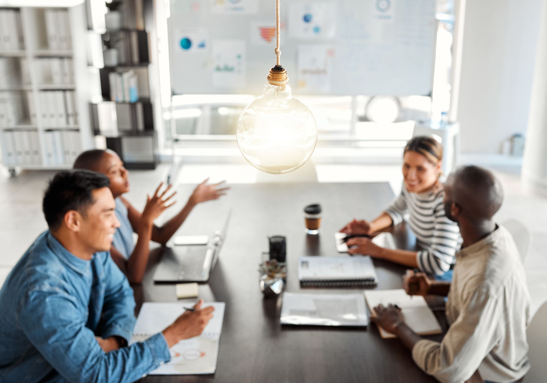 Team meeting around a table in a bright office, with laptops, notes, and a hanging light bulb overhead.