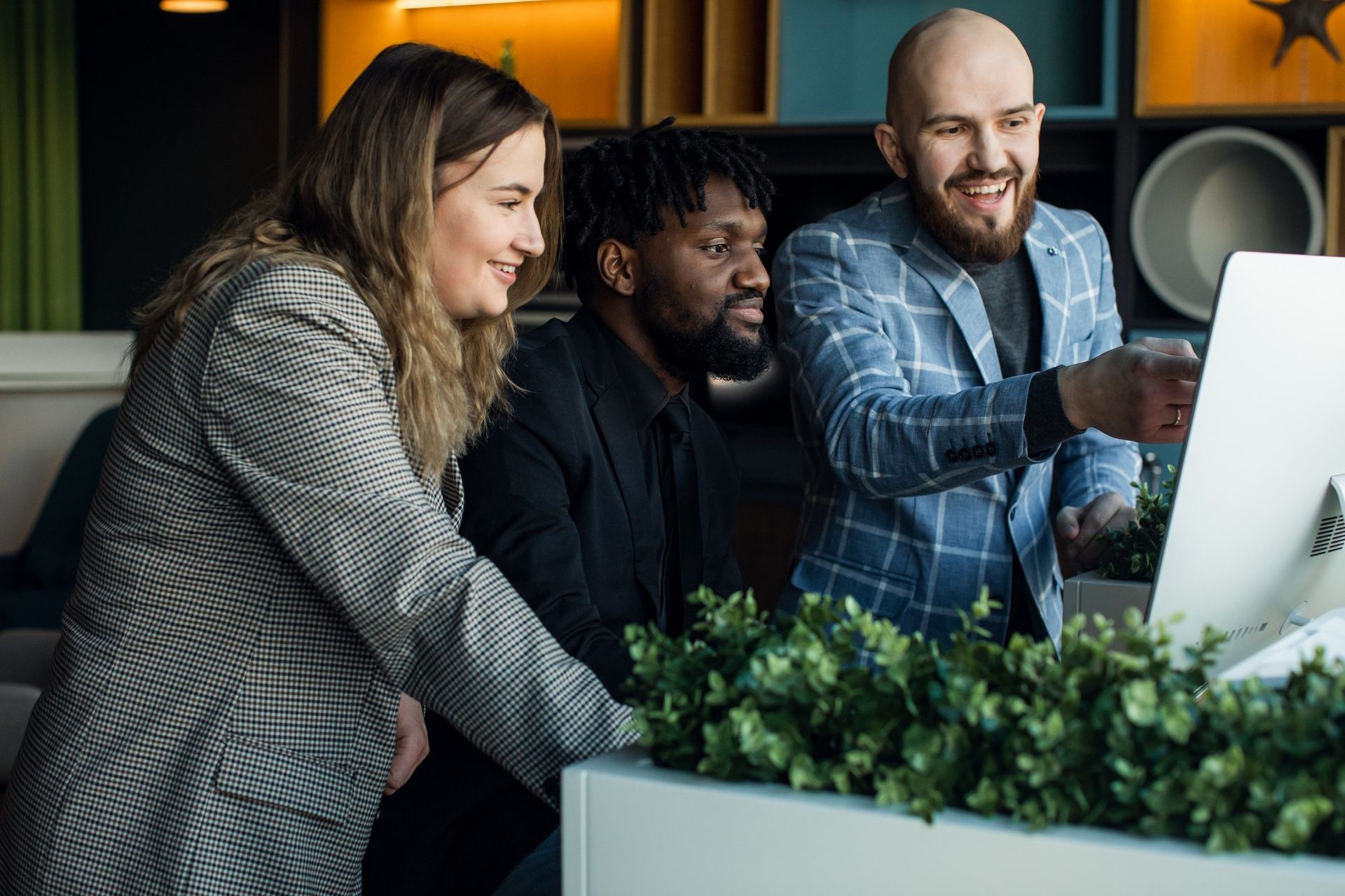 Three coworkers leaning over a desktop monitor in an office, smiling and collaborating.