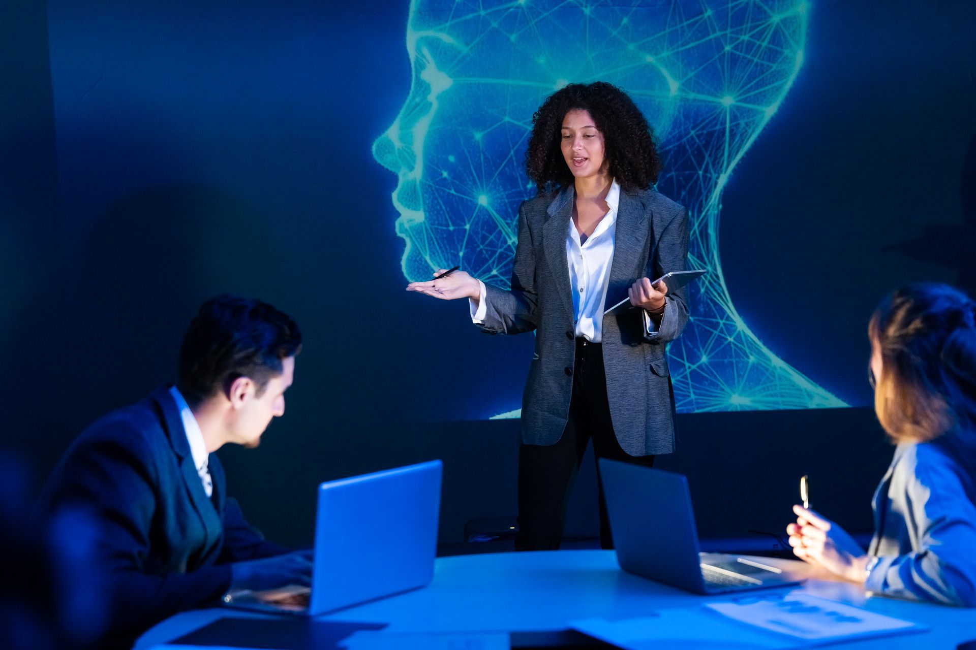 Business meeting with a presenter speaking to two colleagues beside laptops in a blue-lit conference room