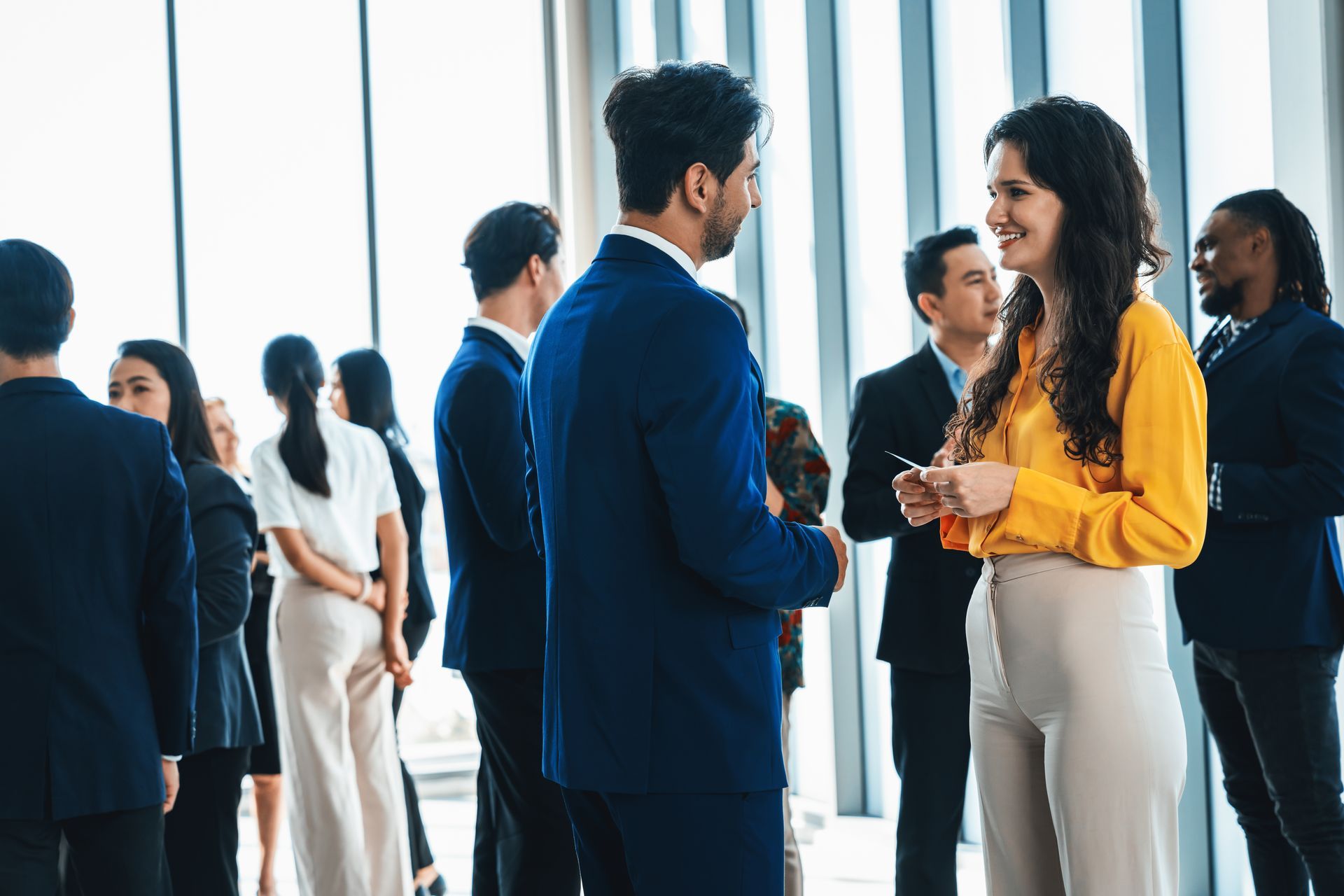 People networking in a bright office, with a woman in a yellow blouse speaking to a man in a blue suit.