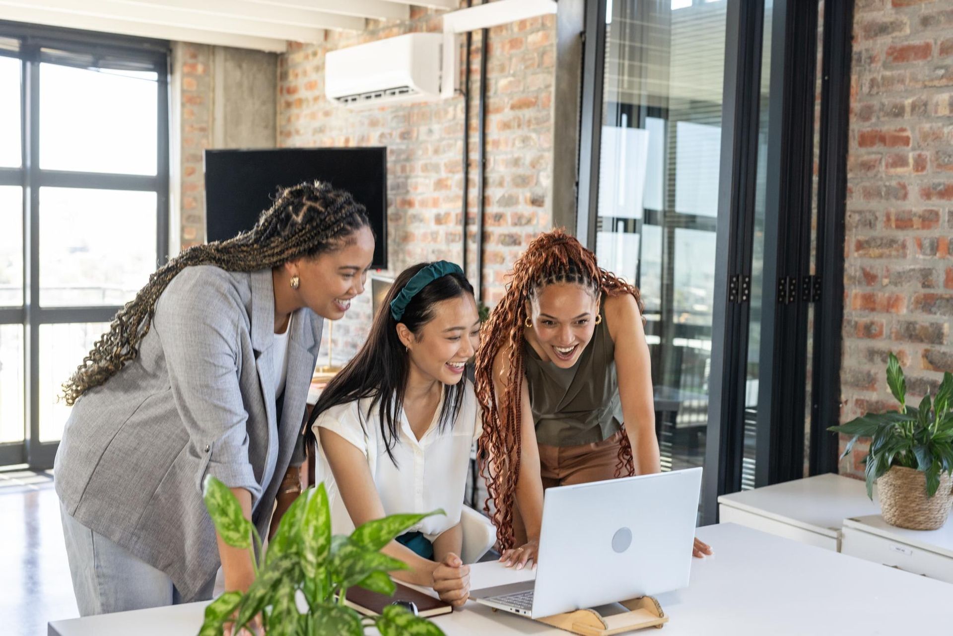 Three coworkers leaning over a laptop in a bright office, smiling and discussing work.