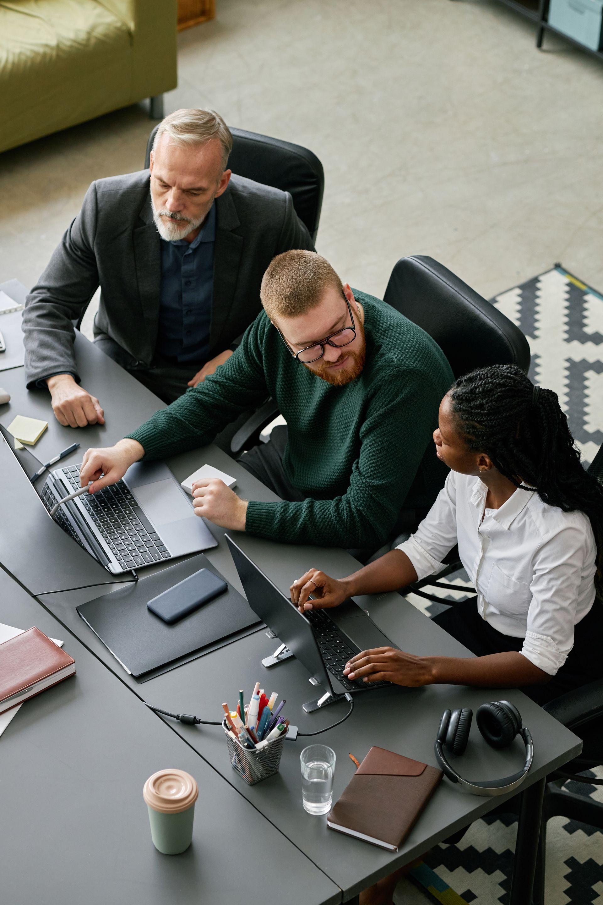 Three coworkers in a meeting around a laptop at a desk with notebooks and coffee cups.