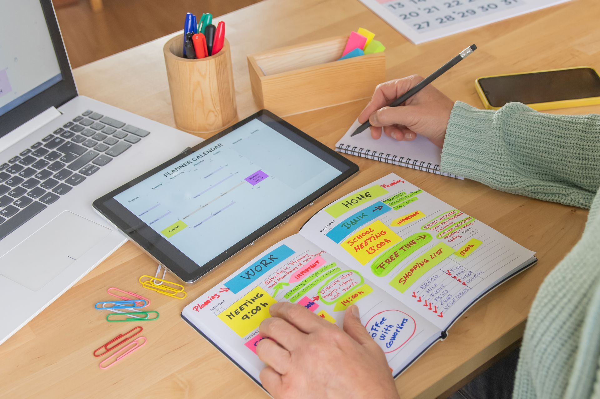 Desk with laptop, tablet, and colorful study notes as someone writes in a notebook