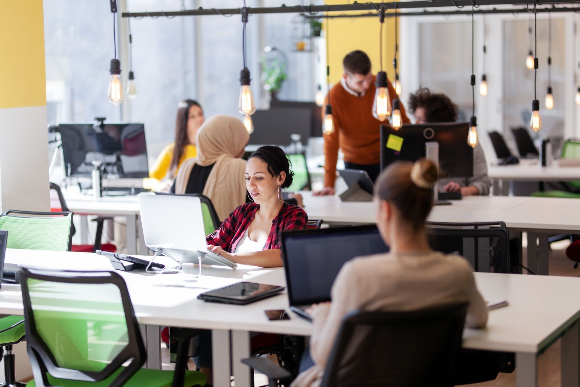 Open-plan office with people working at desks and computers, lit by hanging bulbs