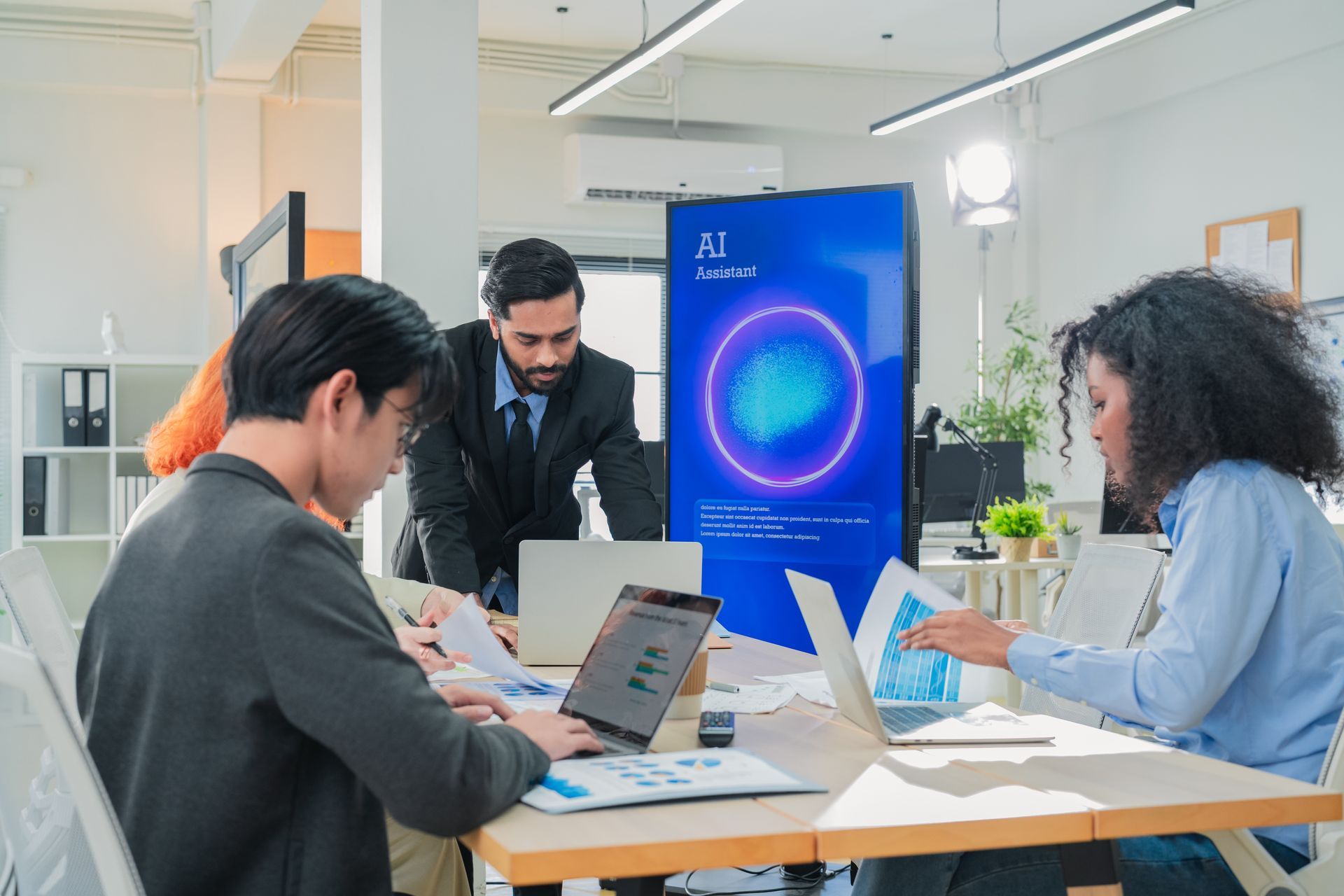 Team collaborating around laptops in a bright office, with a large blue presentation screen in the background.