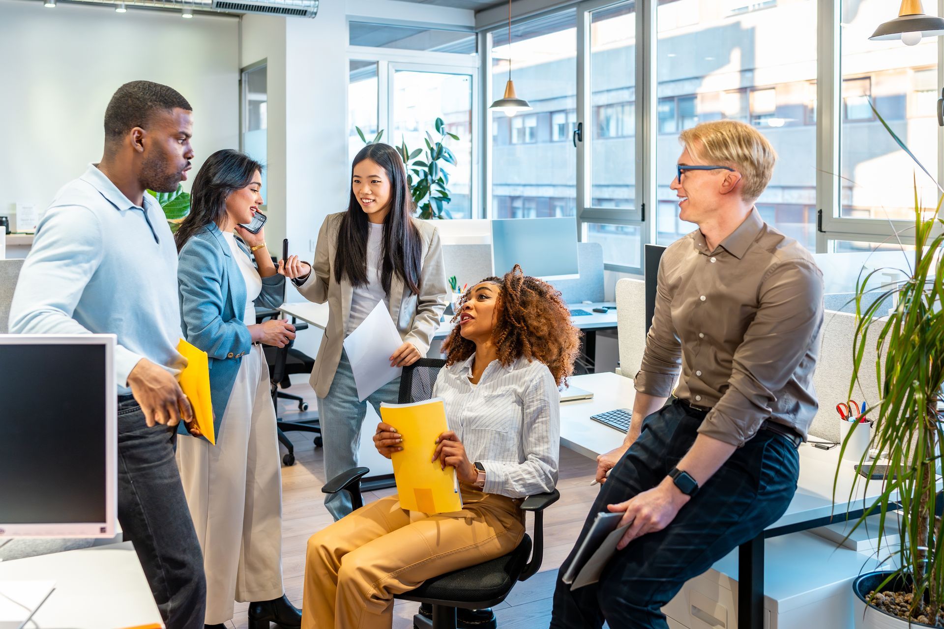Colleagues chatting in a bright modern office lounge, holding papers and a laptop during a casual meeting