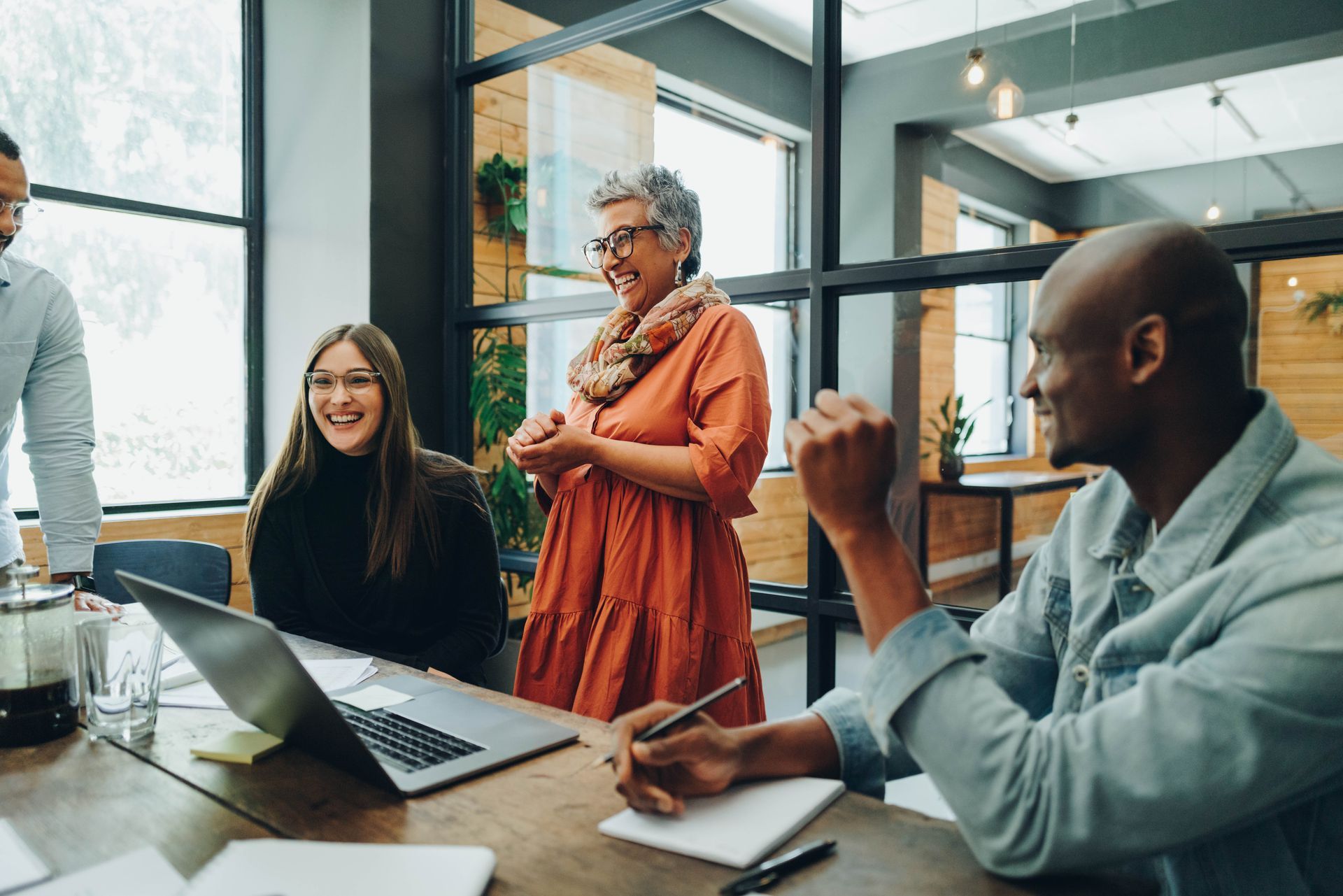 Coworkers in a bright office meeting around a table with laptops and coffee, one person standing and smiling