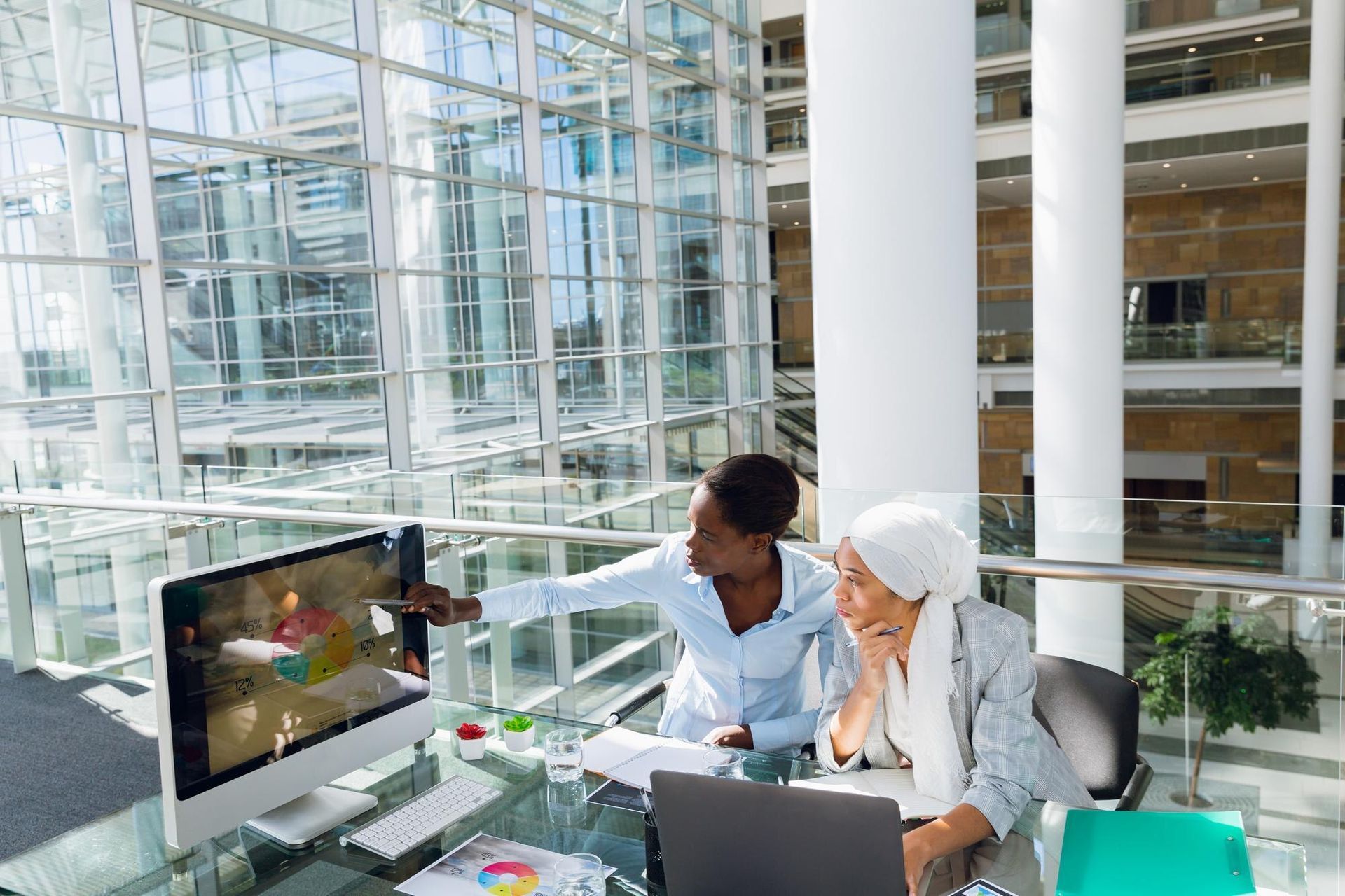 Two coworkers reviewing a laptop in a bright glass atrium, with a monitor showing a video on the table.