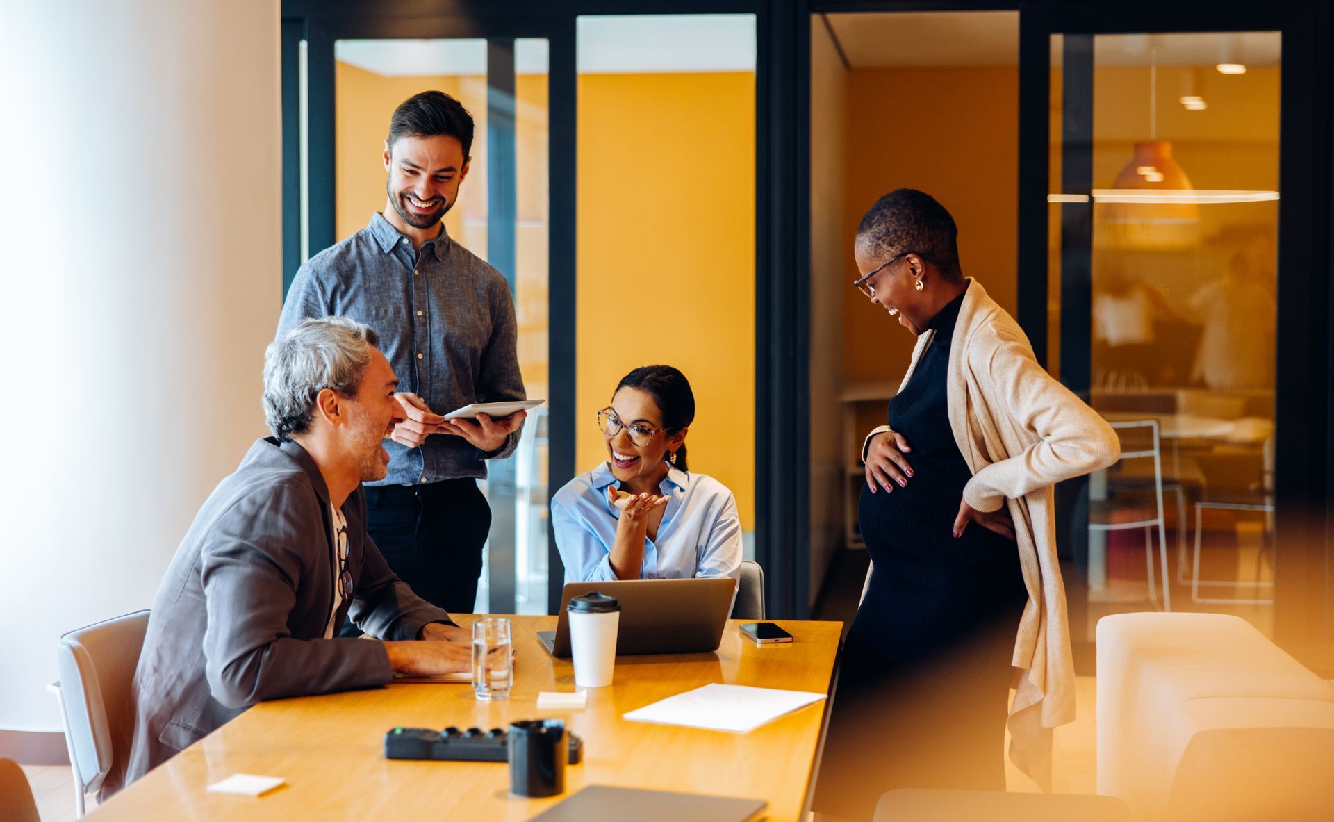 Five people in a modern conference room discussing around a table with laptops and coffee mugs.