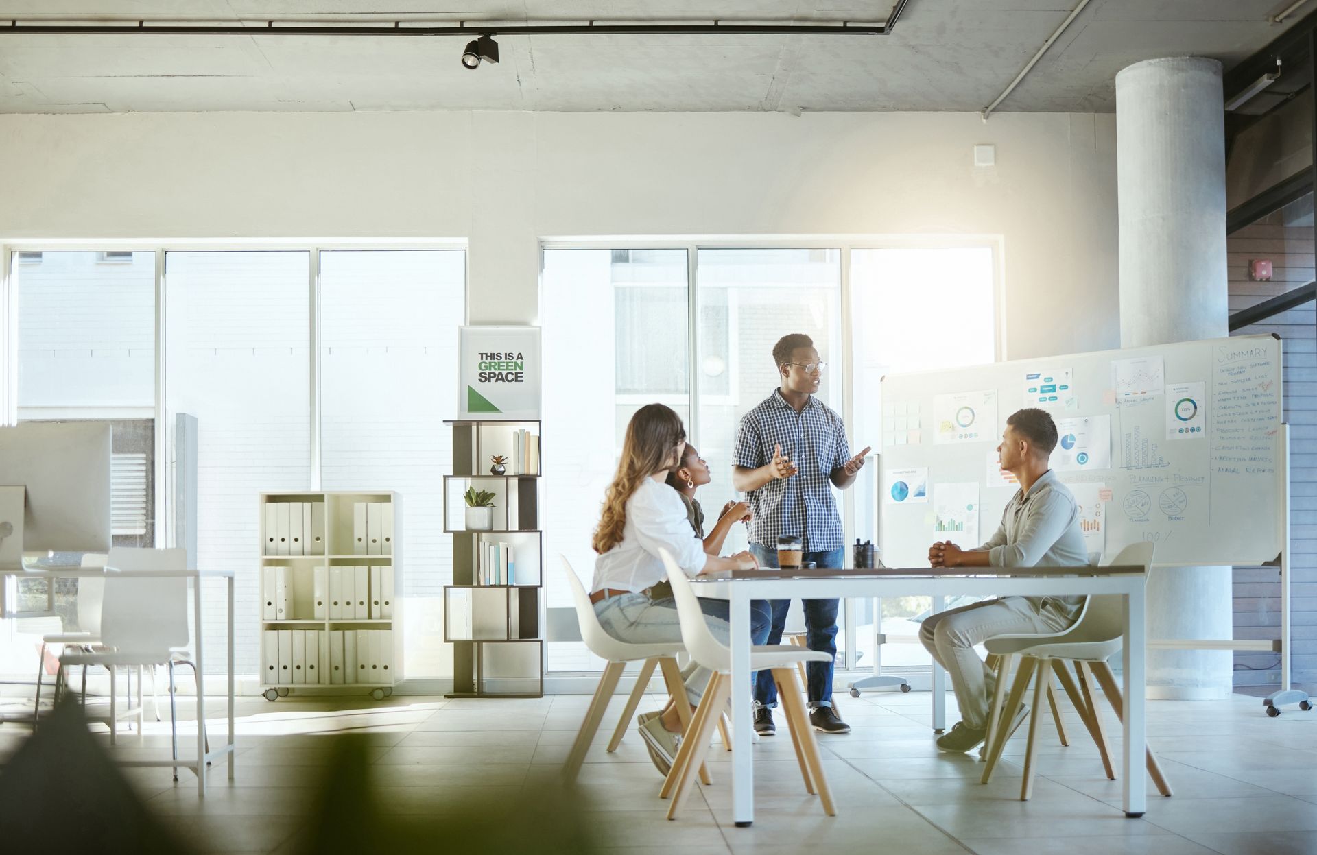 Three coworkers meeting around a table in a bright office with a whiteboard and cabinets.