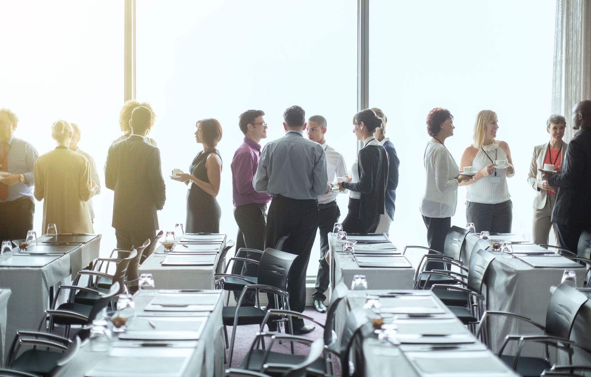 People standing and talking in a bright conference room with rows of tables and chairs