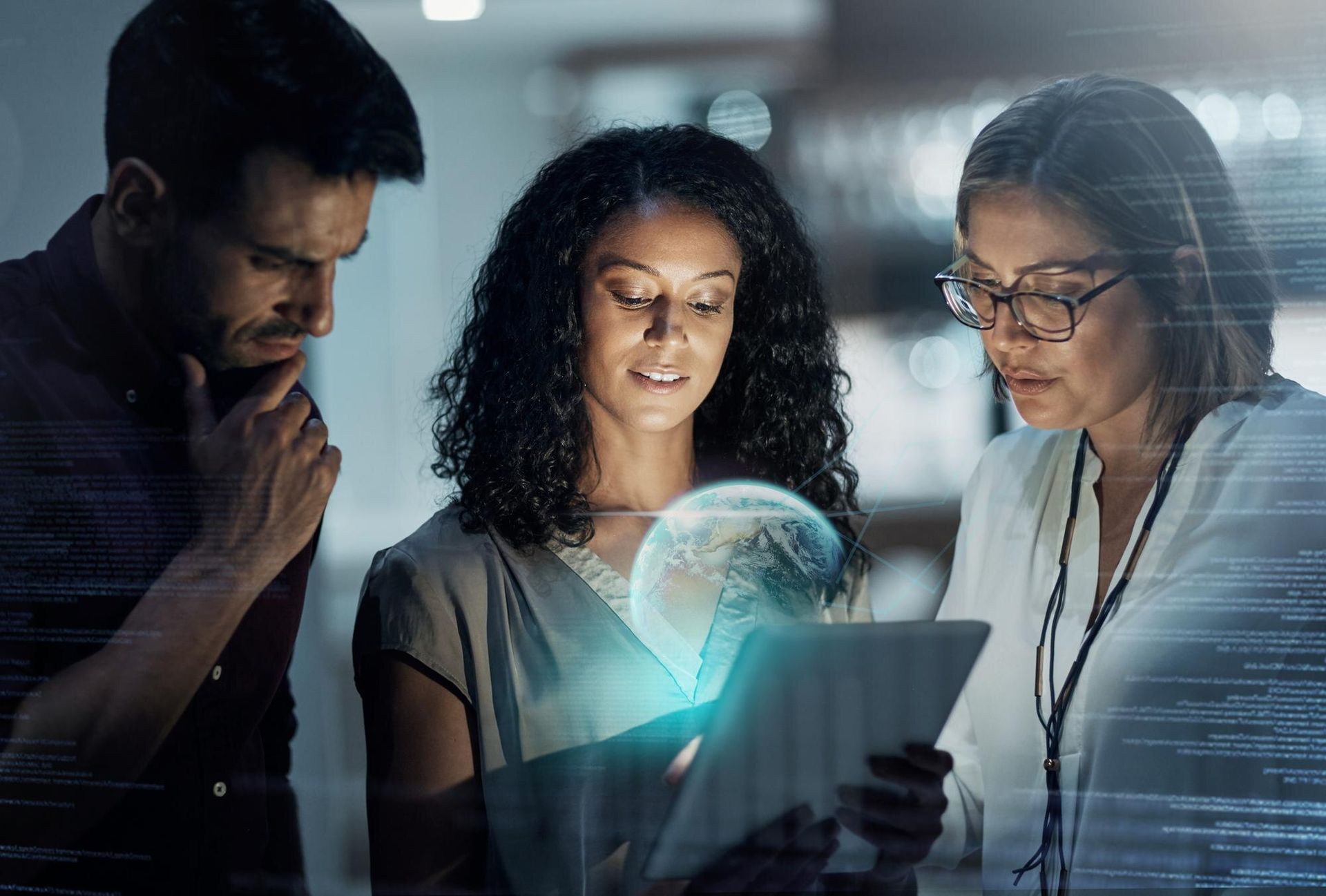 Three coworkers study a glowing tablet in a dark office, focused on a holographic interface.