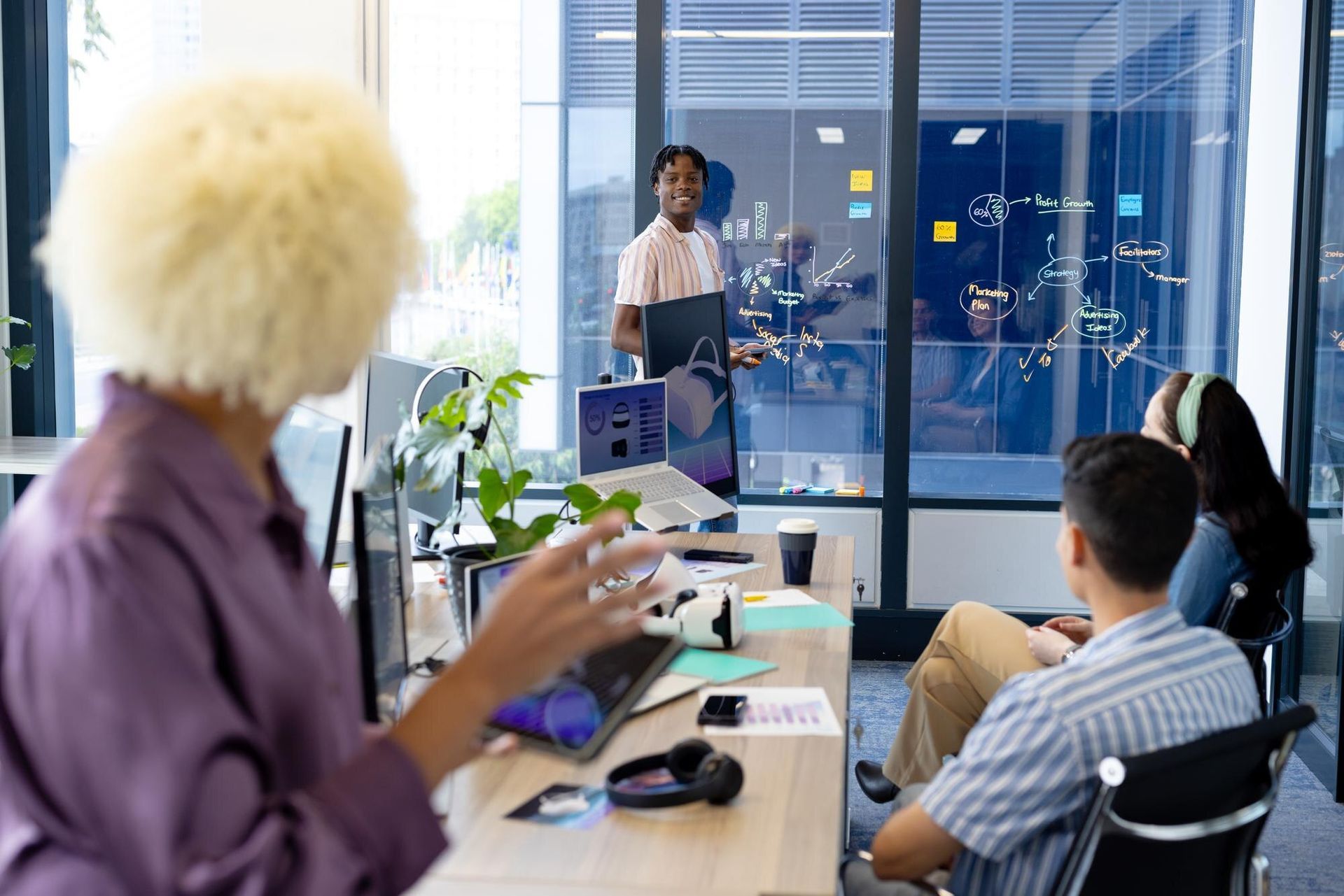 Office meeting with a presenter speaking to seated coworkers in a glass-walled conference room