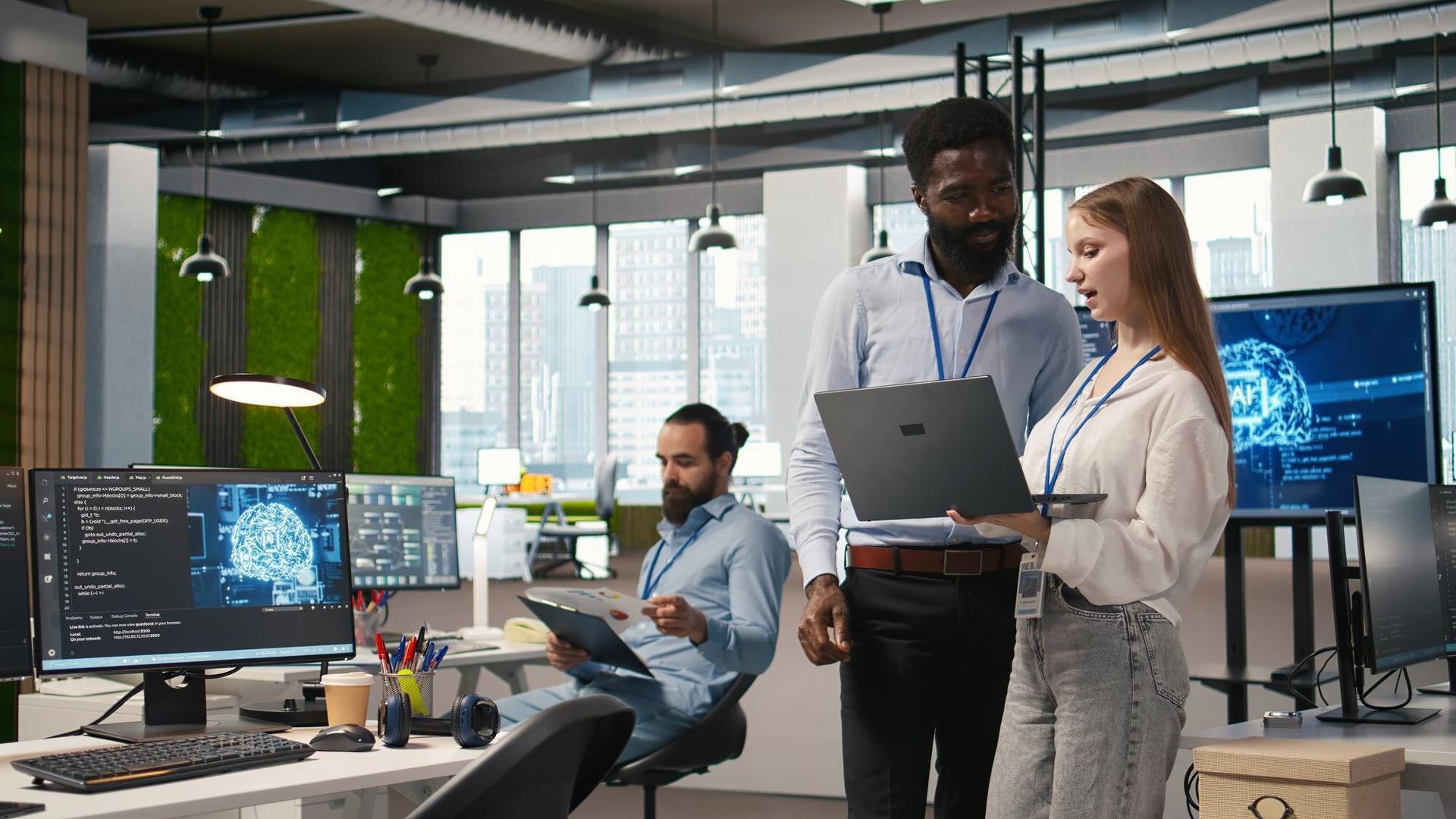 Coworkers in a modern office reviewing data on laptops and monitors near a table with plants and hanging lights