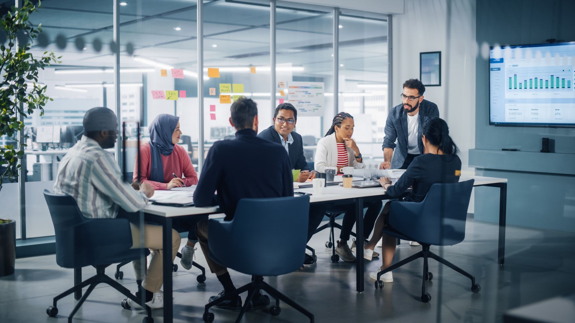 Business team meeting in a glass conference room, with charts on a wall screen.