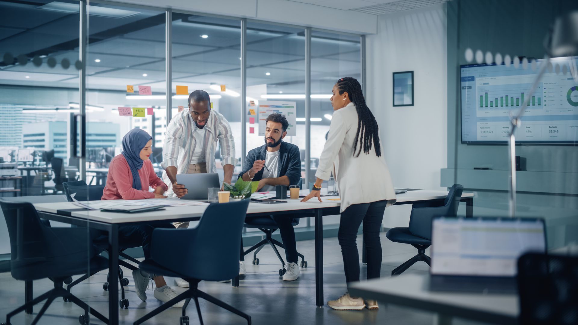 Team meeting in a modern office, with four people gathered around a table and a presentation screen nearby.