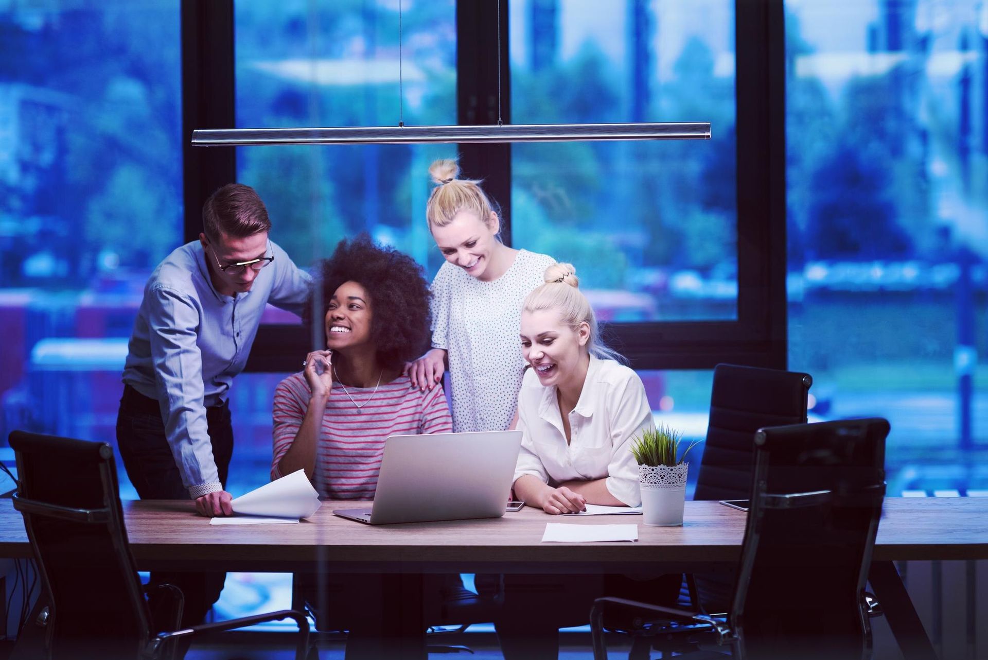 Four coworkers gather around a laptop in a modern office, leaning in and discussing work.