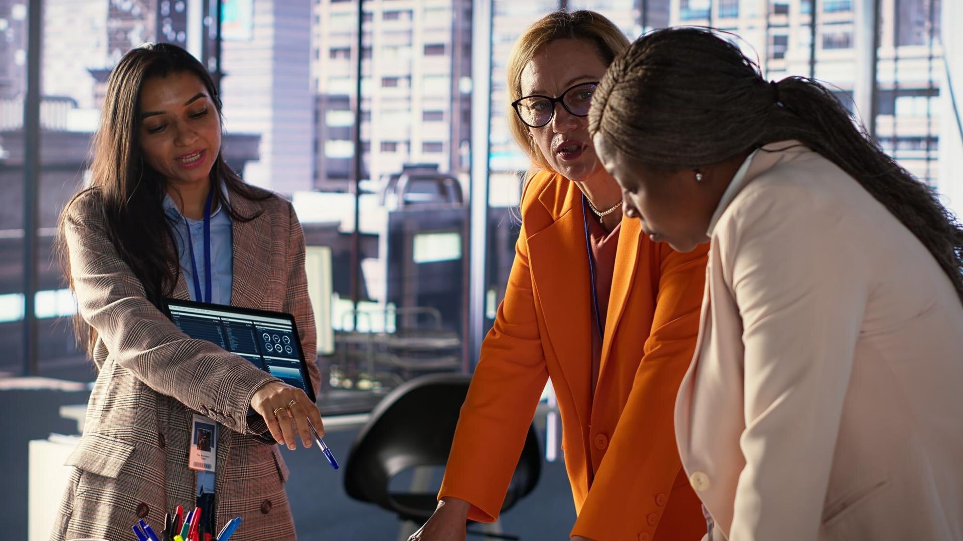 Three coworkers lean over a table, pointing at papers in a bright office meeting room.