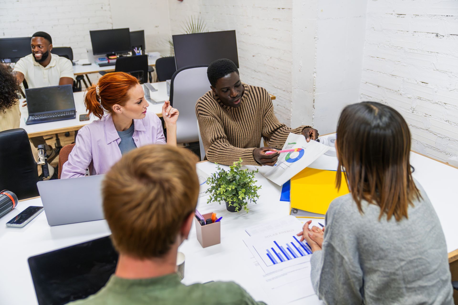 Team meeting around a table with laptops, charts, and a plant in a bright office