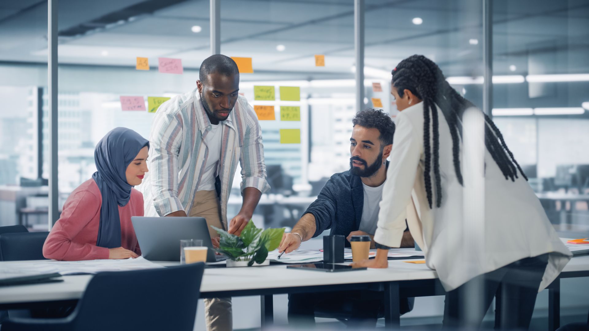 Coworkers gathered around a laptop in a bright modern office, discussing documents at a table.