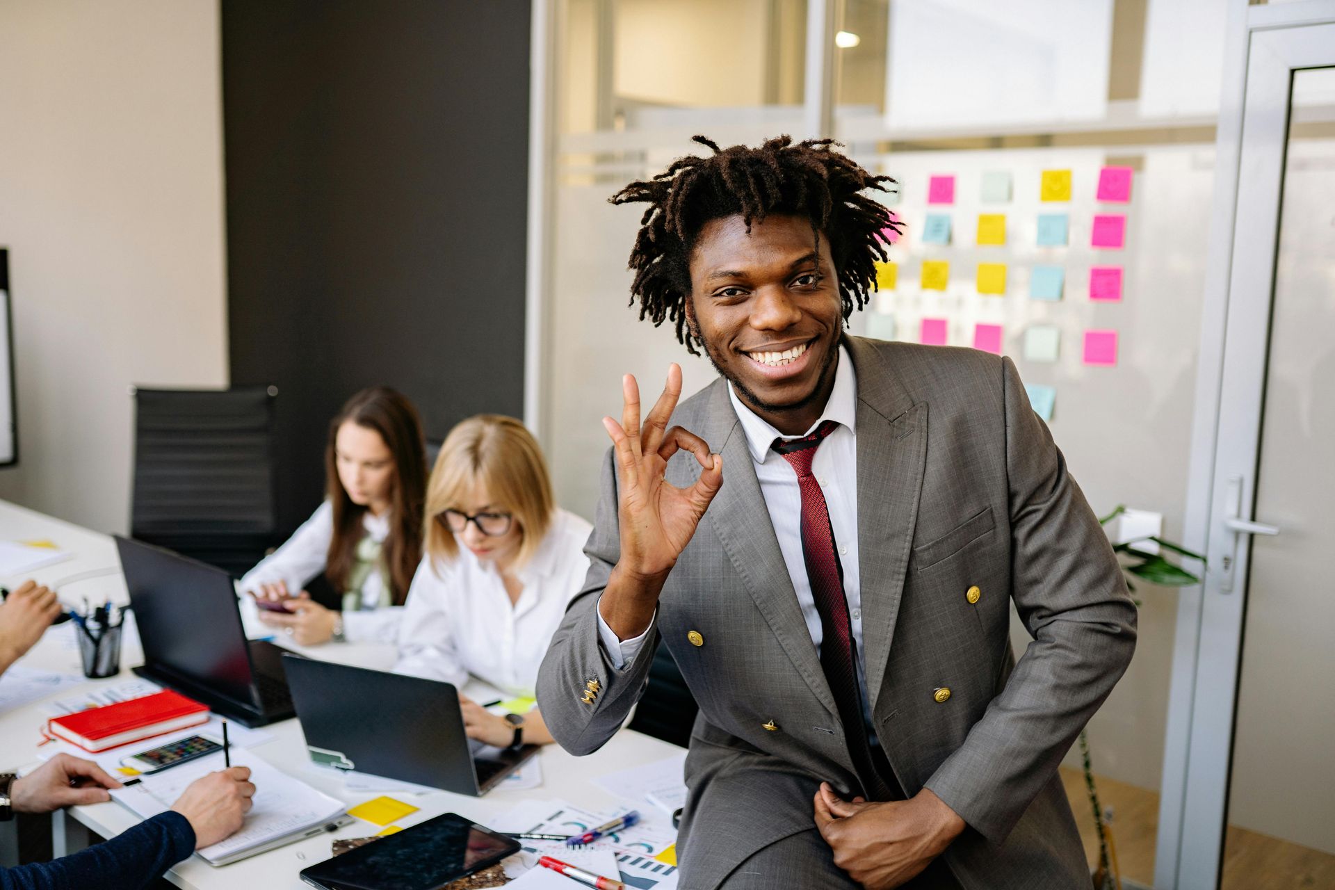 Smiling man in a gray suit poses in an office while coworkers work at laptops behind him