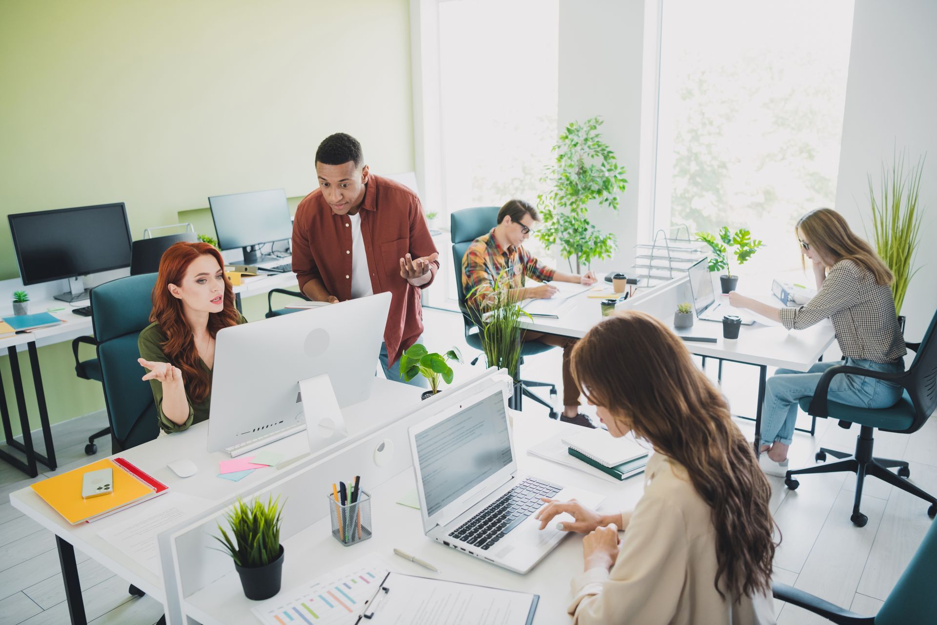 Office team collaborating around computers in a bright, modern workspace with white desks and plants