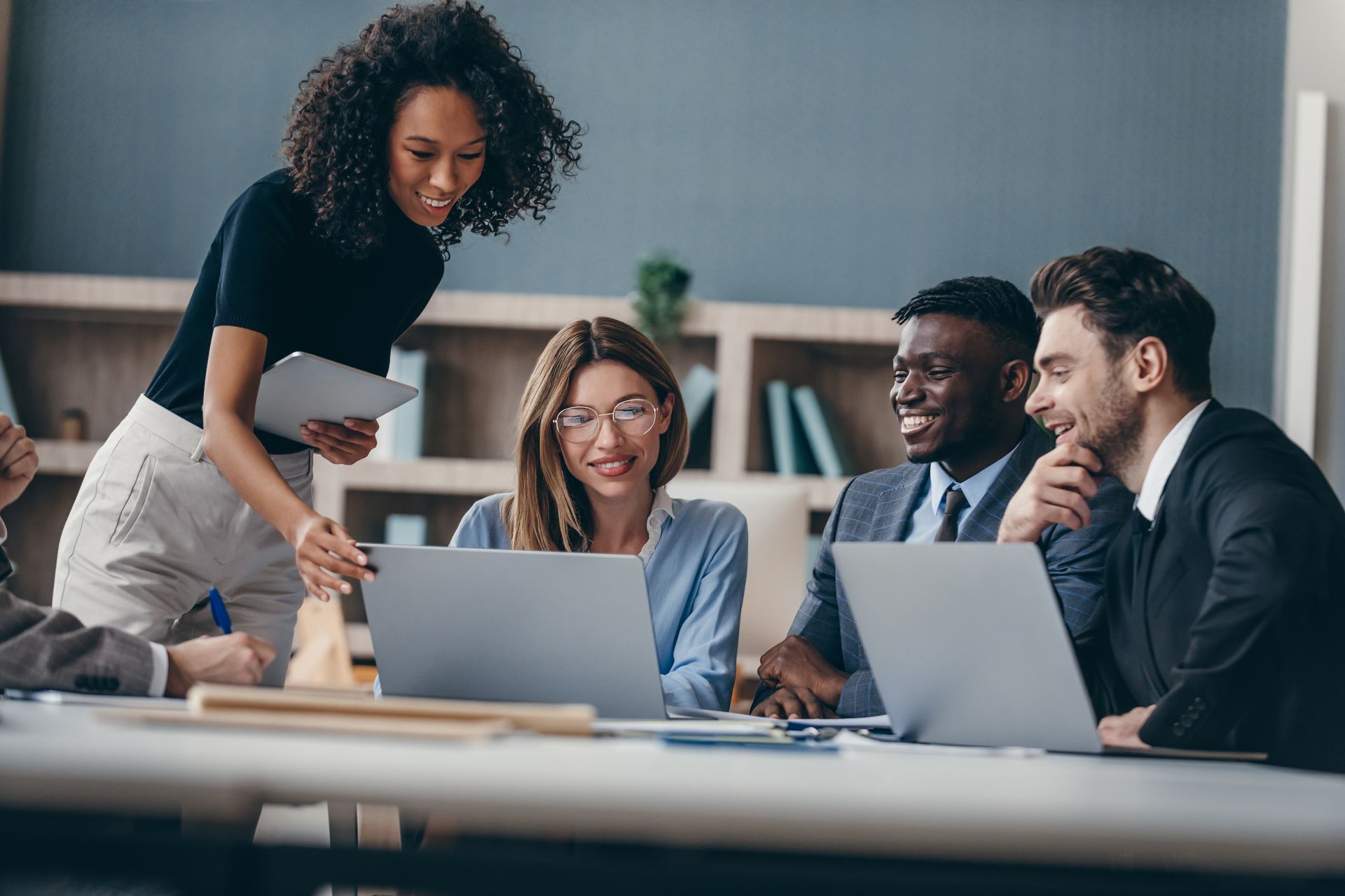 Team meeting in a modern office, with four colleagues around laptops and one woman standing and smiling.