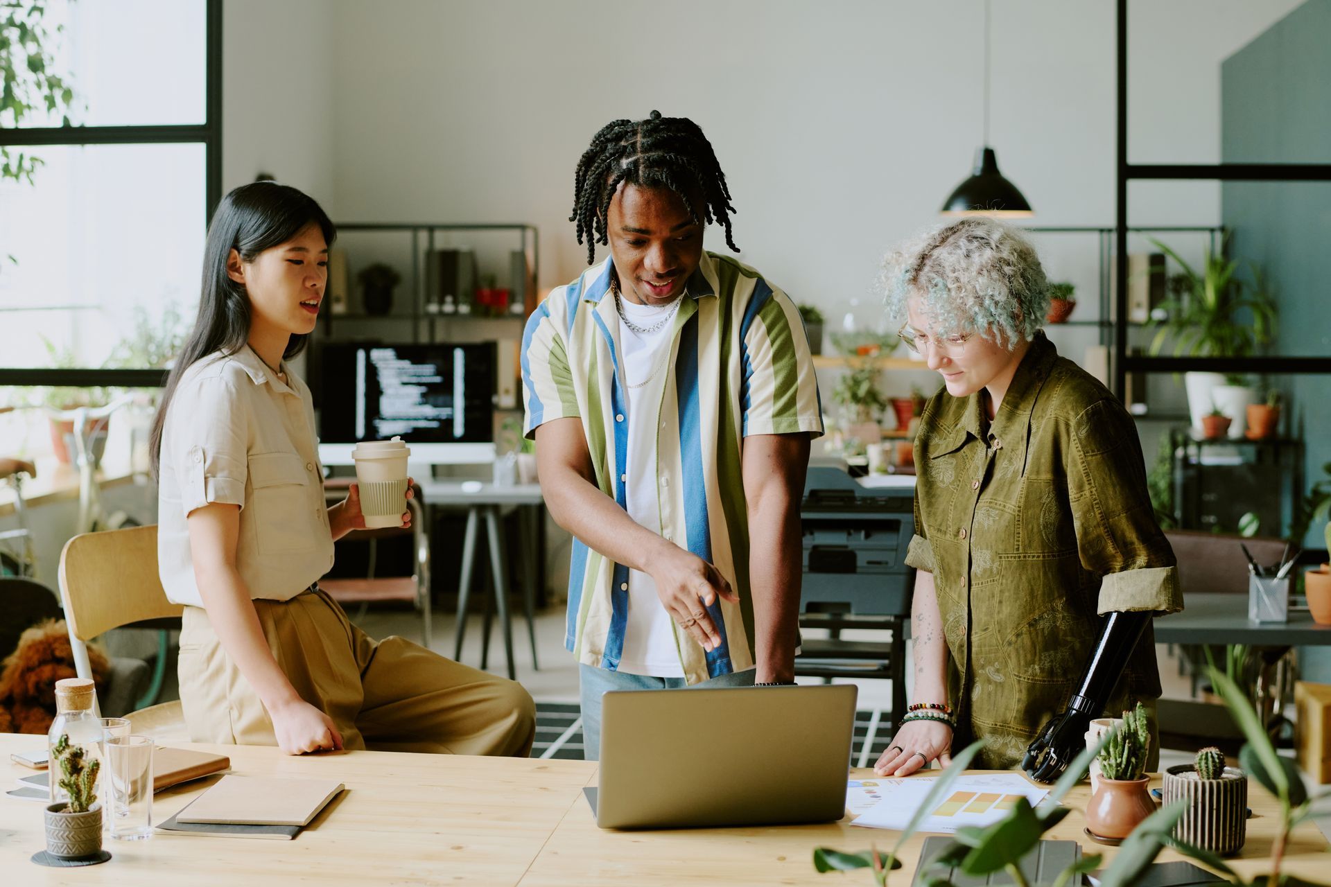 Three coworkers stand around a laptop in a bright office, discussing work at a table.