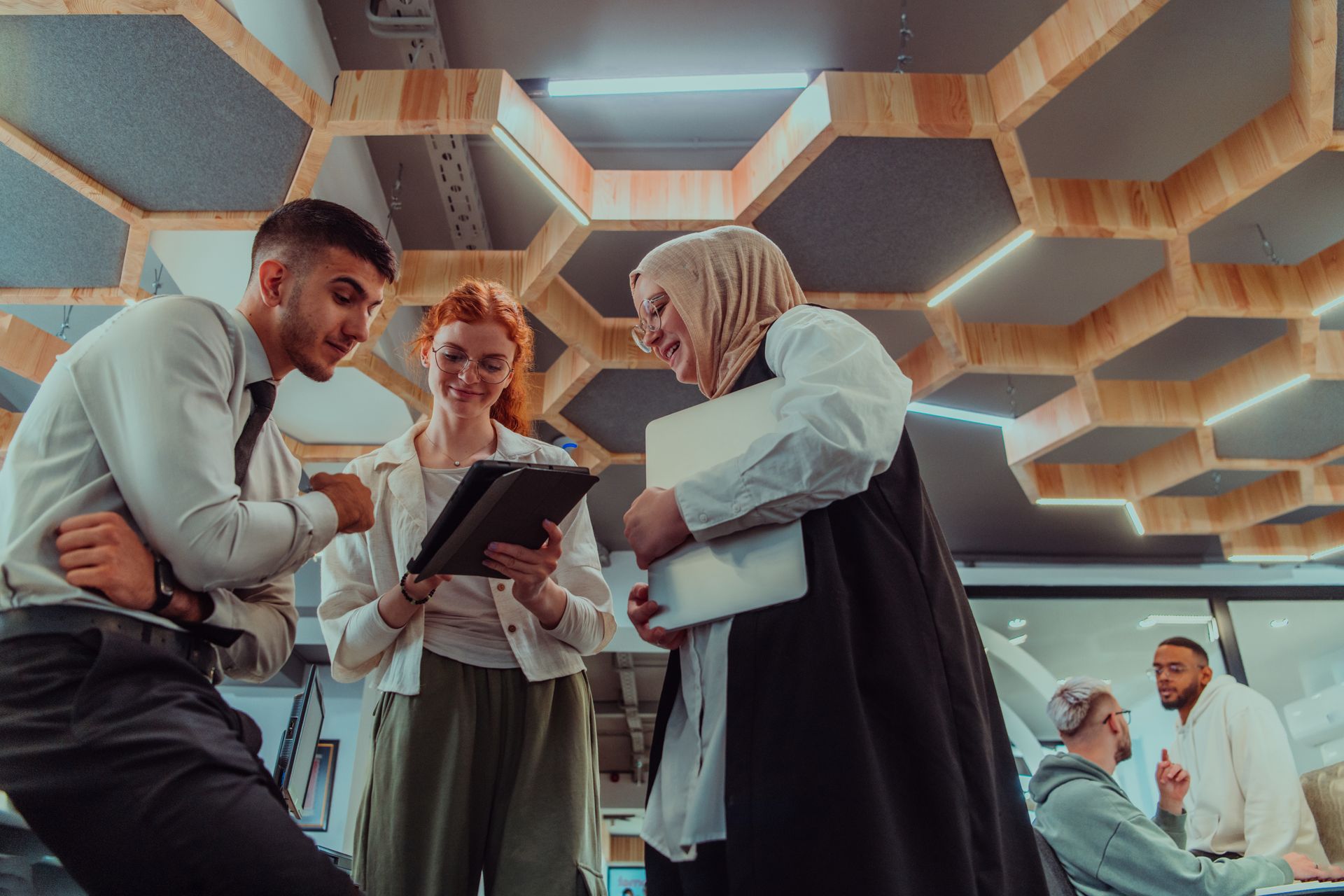 People collaborating in a modern office, discussing a tablet under hexagonal ceiling lights.