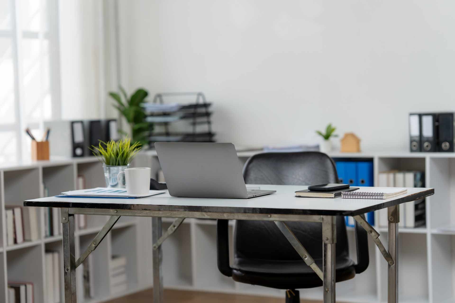 Office desk with laptop, plant, and papers. Black chair, white walls, and bookshelves in the background.