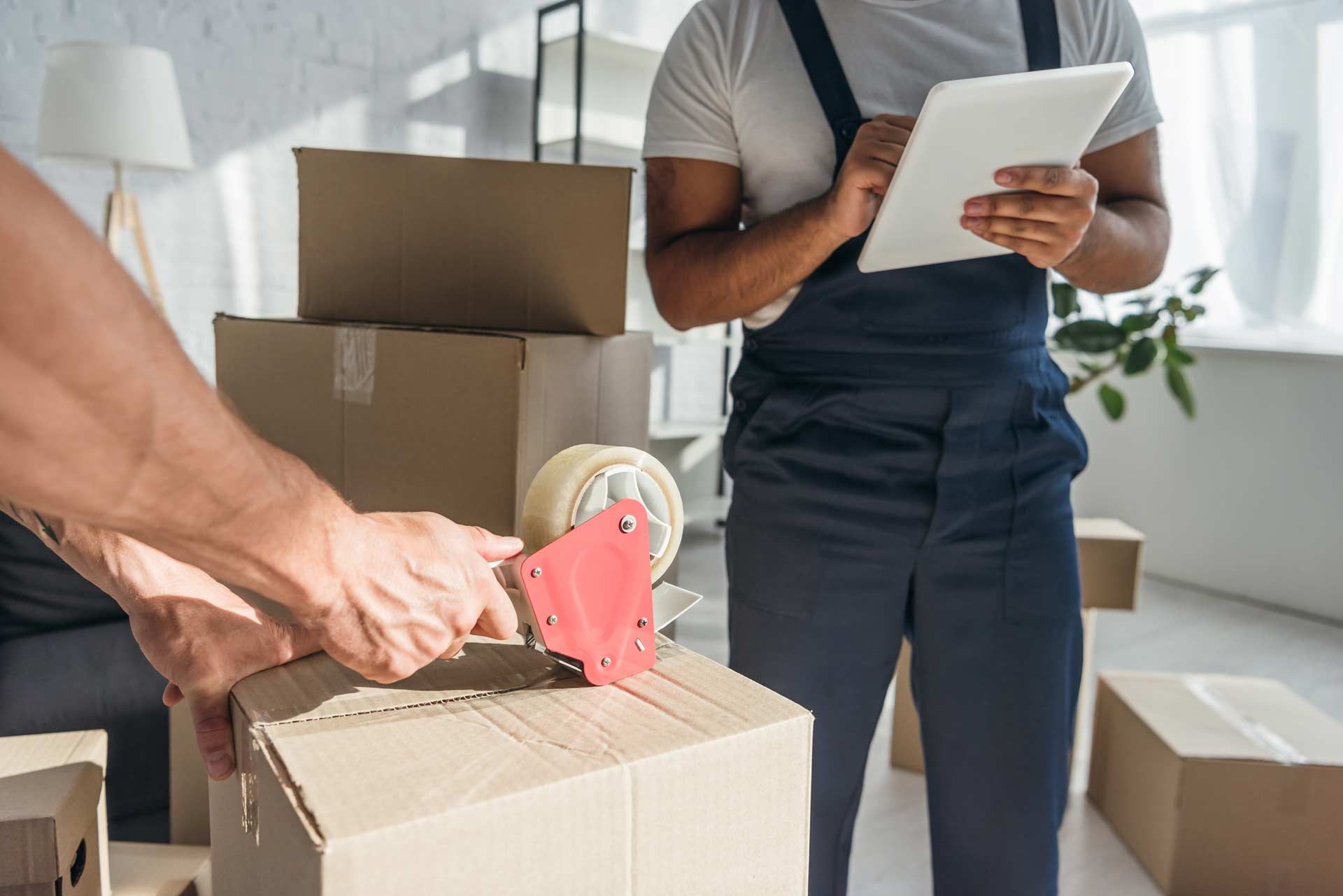 Movers packing boxes, one taping, the other using a tablet indoors.