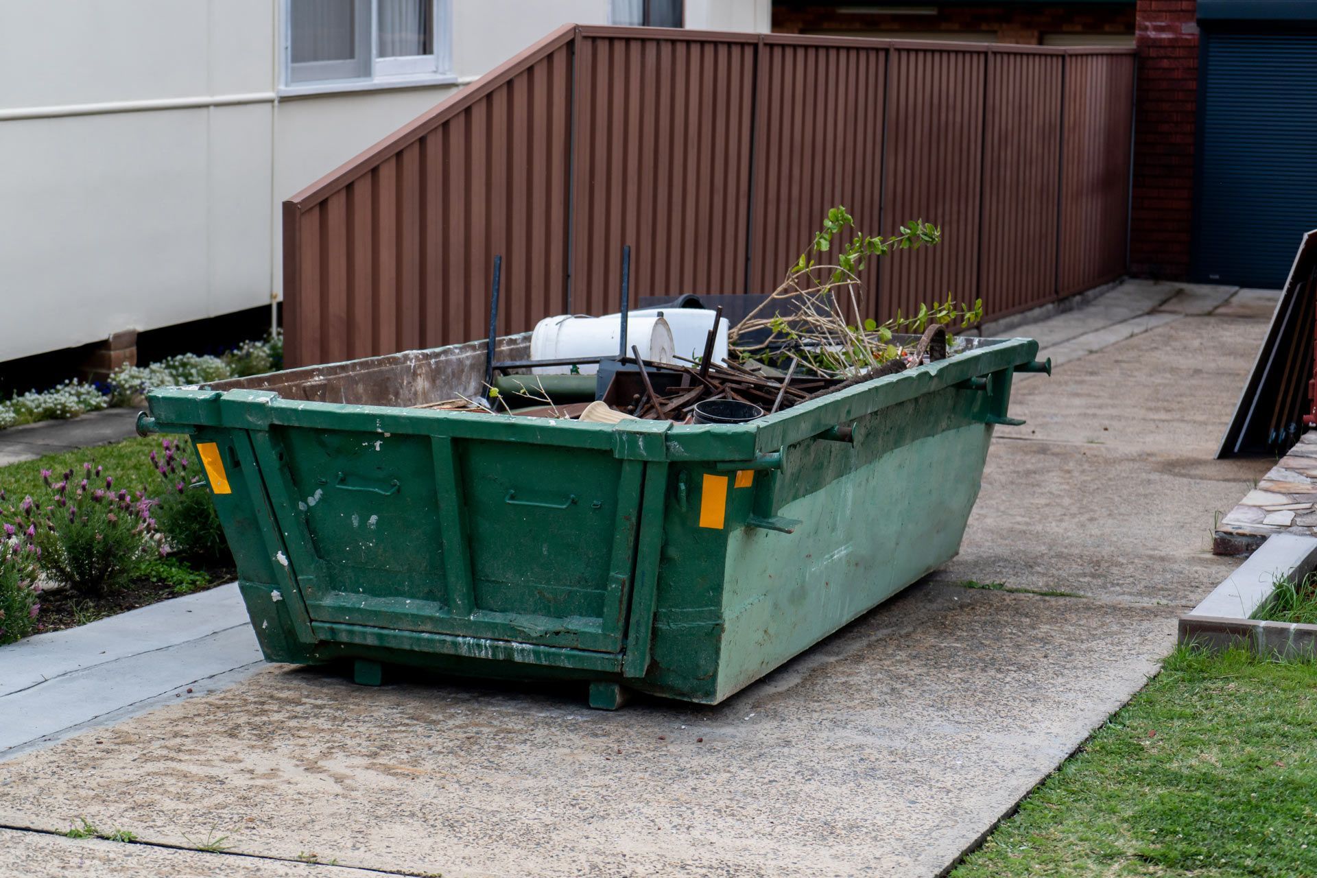 Green dumpster on a concrete driveway, filled with yard waste, next to a brown fence.
